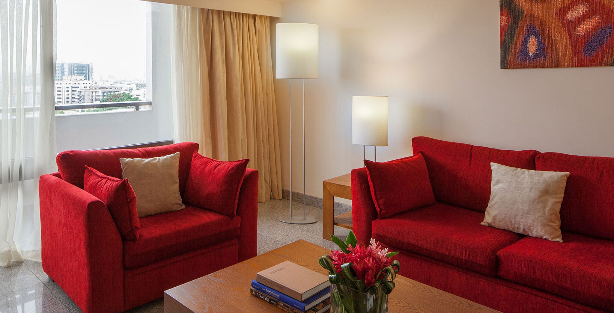 A hotel room with two red sofas, a small wooden table with books and a vase of flowers, a floor lamp, and a painting on the wall.