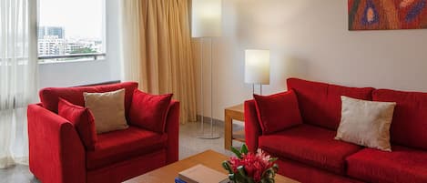Una habitación de hotel con dos sofás rojos, una pequeña mesa de madera con libros y un jarrón de flores, una lámpara de pie y un cuadro en la pared.