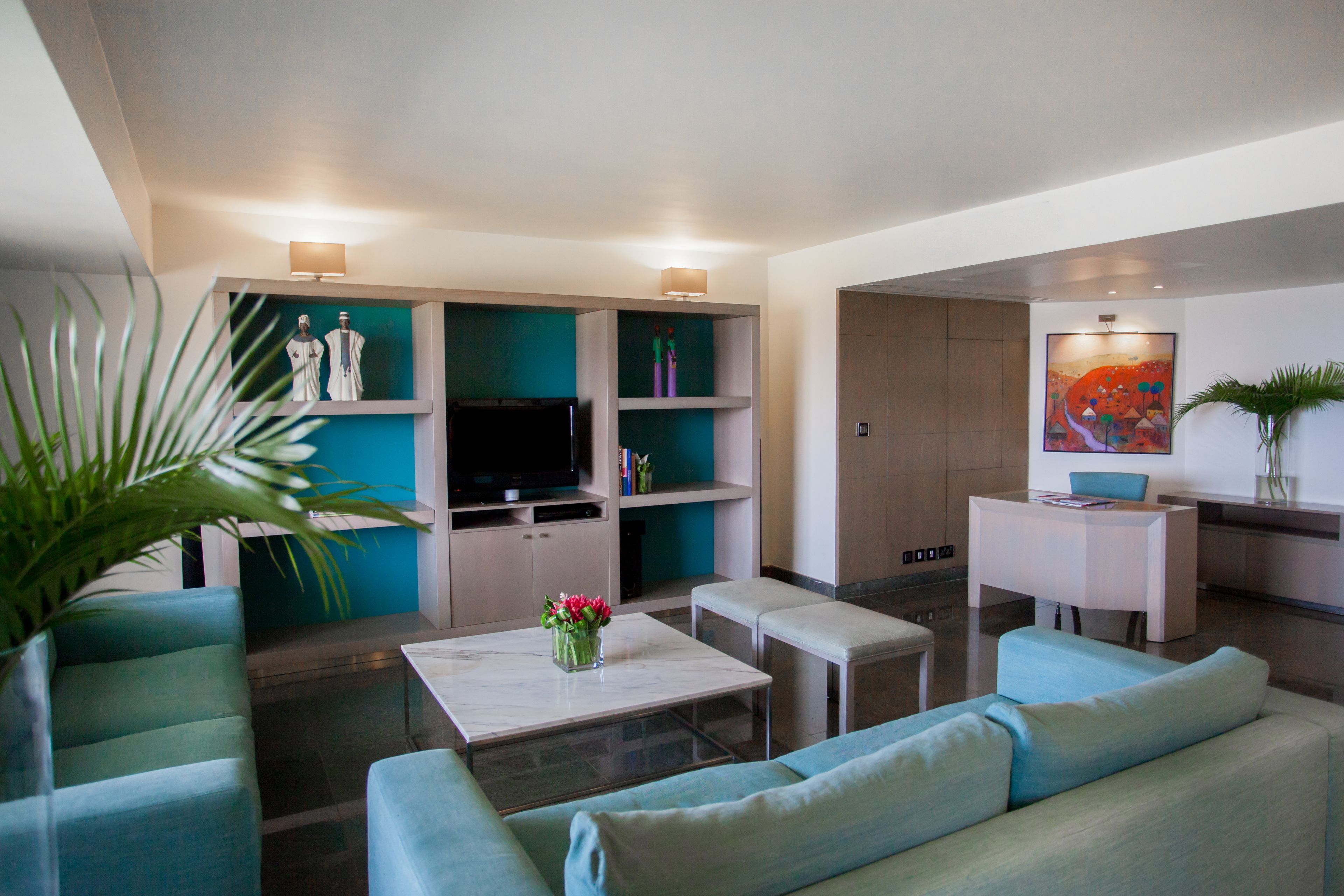 A modern living room with a sofa, coffee table, and TV. There are shelves with decorative items and a desk with a chair.