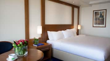 A hotel room with a large bed, a bedside table with a lamp, a small table with a cup and flowers, and a painting on the wall.