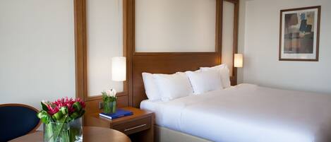 A hotel room with a large bed, a bedside table with a lamp, a small table with a cup and flowers, and a painting on the wall.