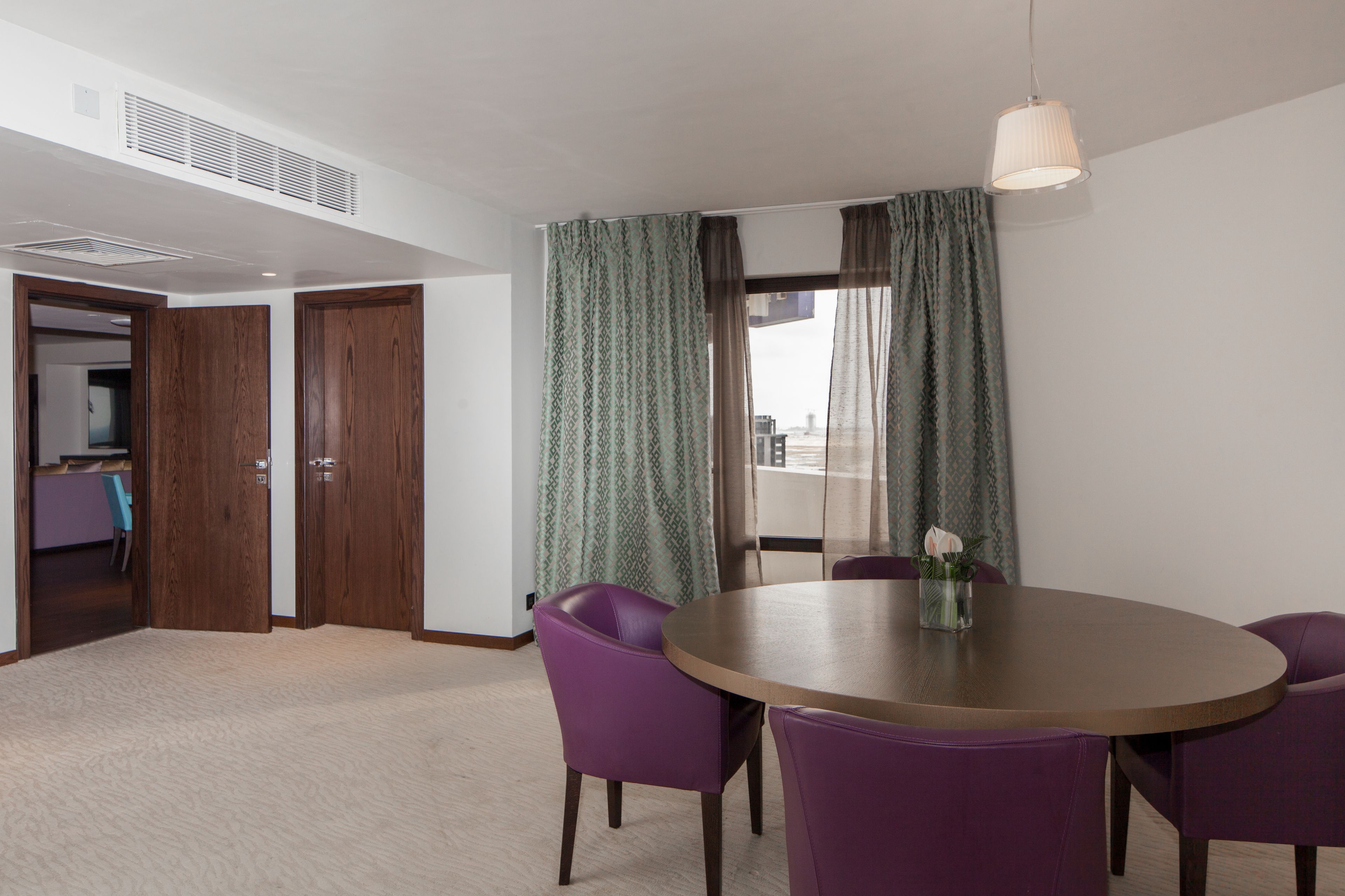 A room with a round table, purple chairs, a hanging light fixture, and a view through a window with curtains.
