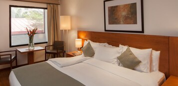 Habitación de hotel con una cama bien tendida, un escritorio con silla, una lámpara y un cuadro enmarcado en la pared.