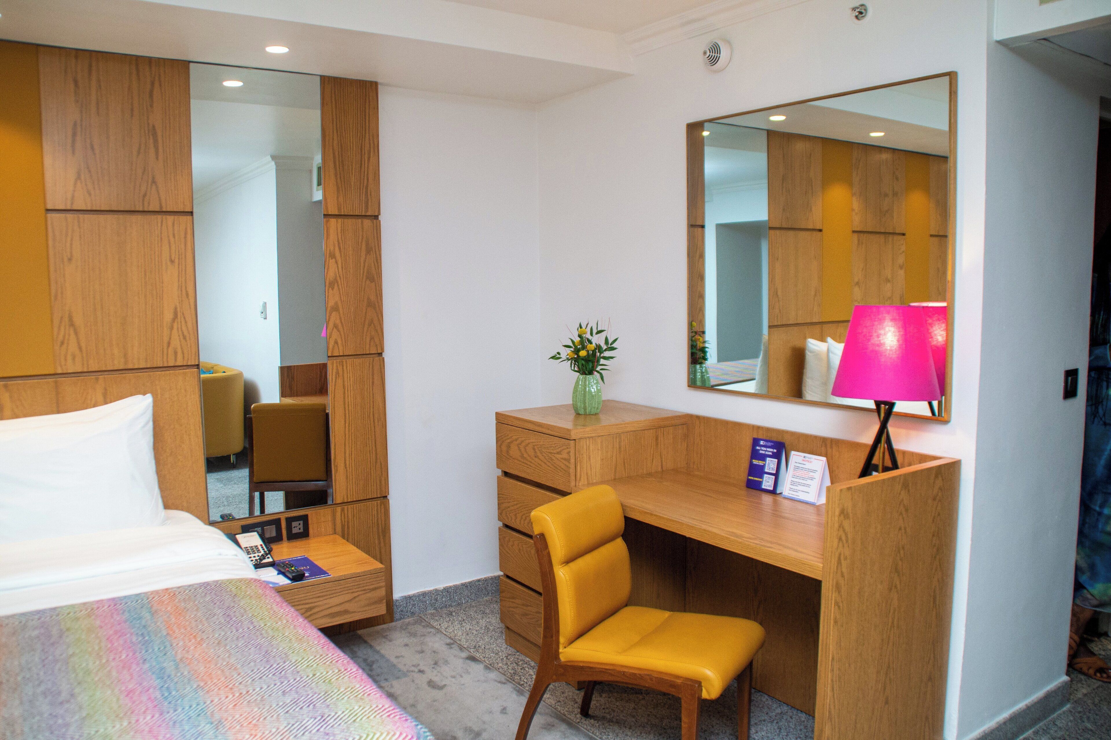 A hotel room with a bed, a desk, a chair, a mirror, and a lamp.