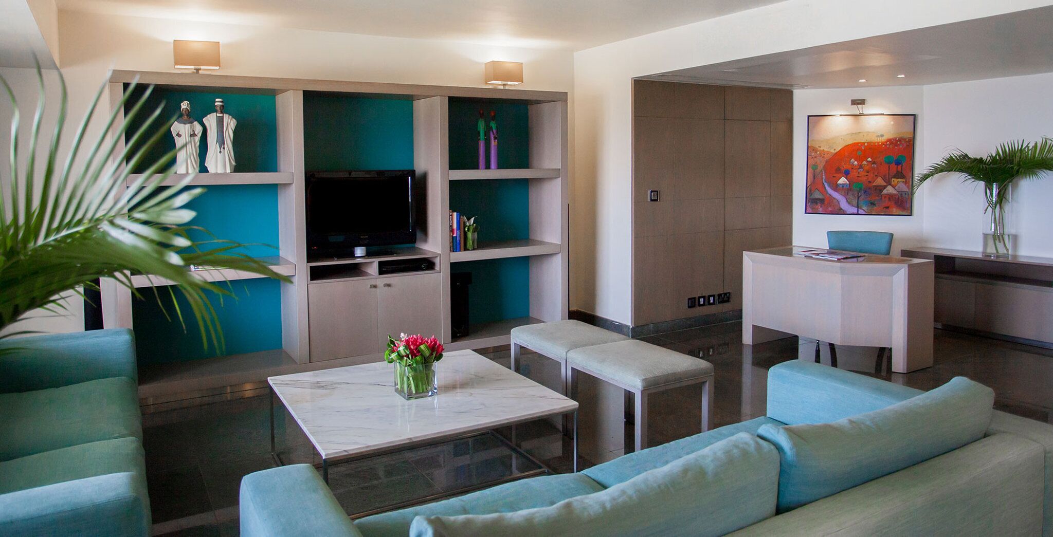 A modern living room with teal sofas, a coffee table with a vase of flowers, a flat-screen TV, and a bookshelf.