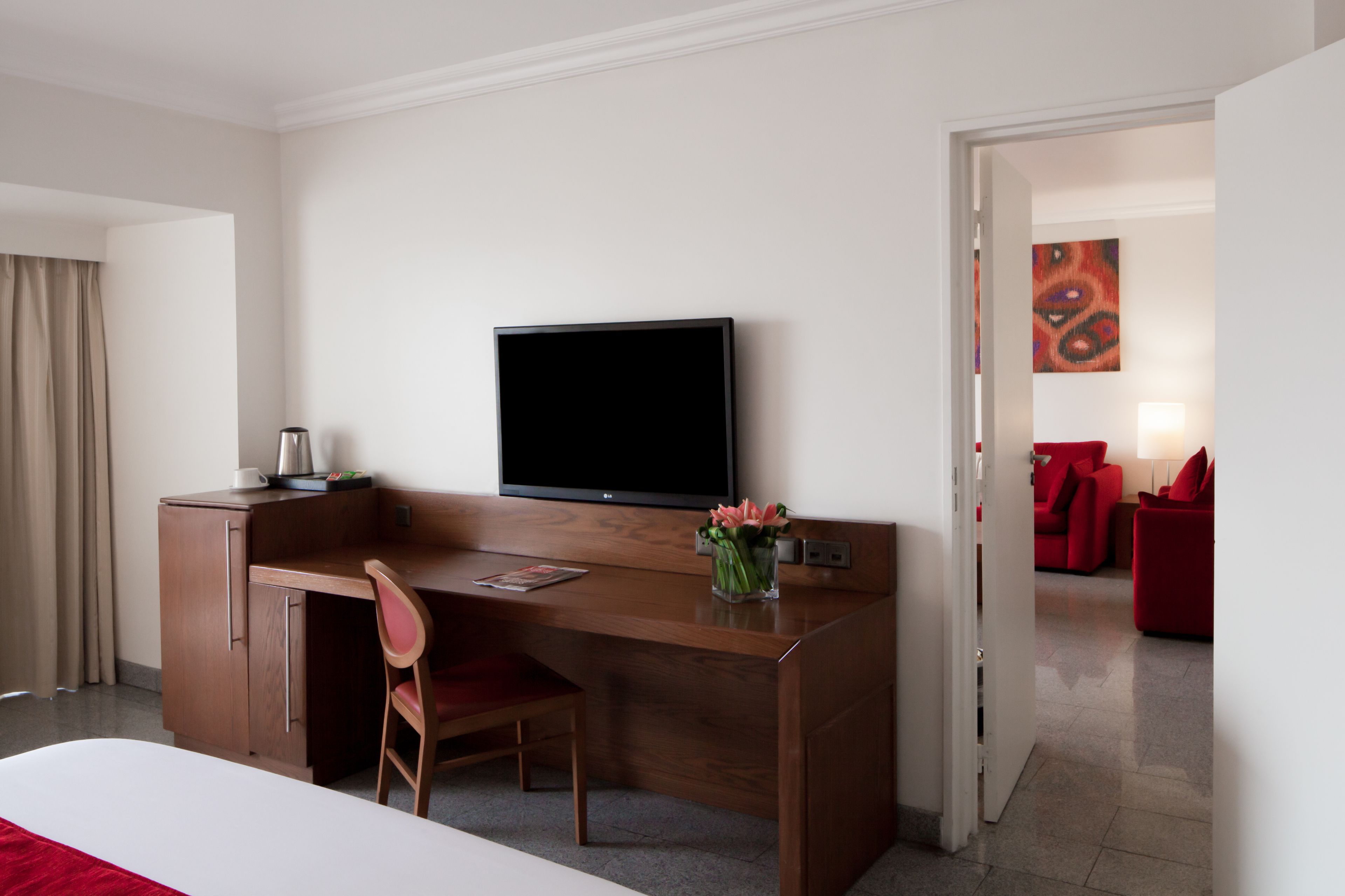 A hotel room with a wooden desk, a television, a chair, a bed, and a red sofa in the background.