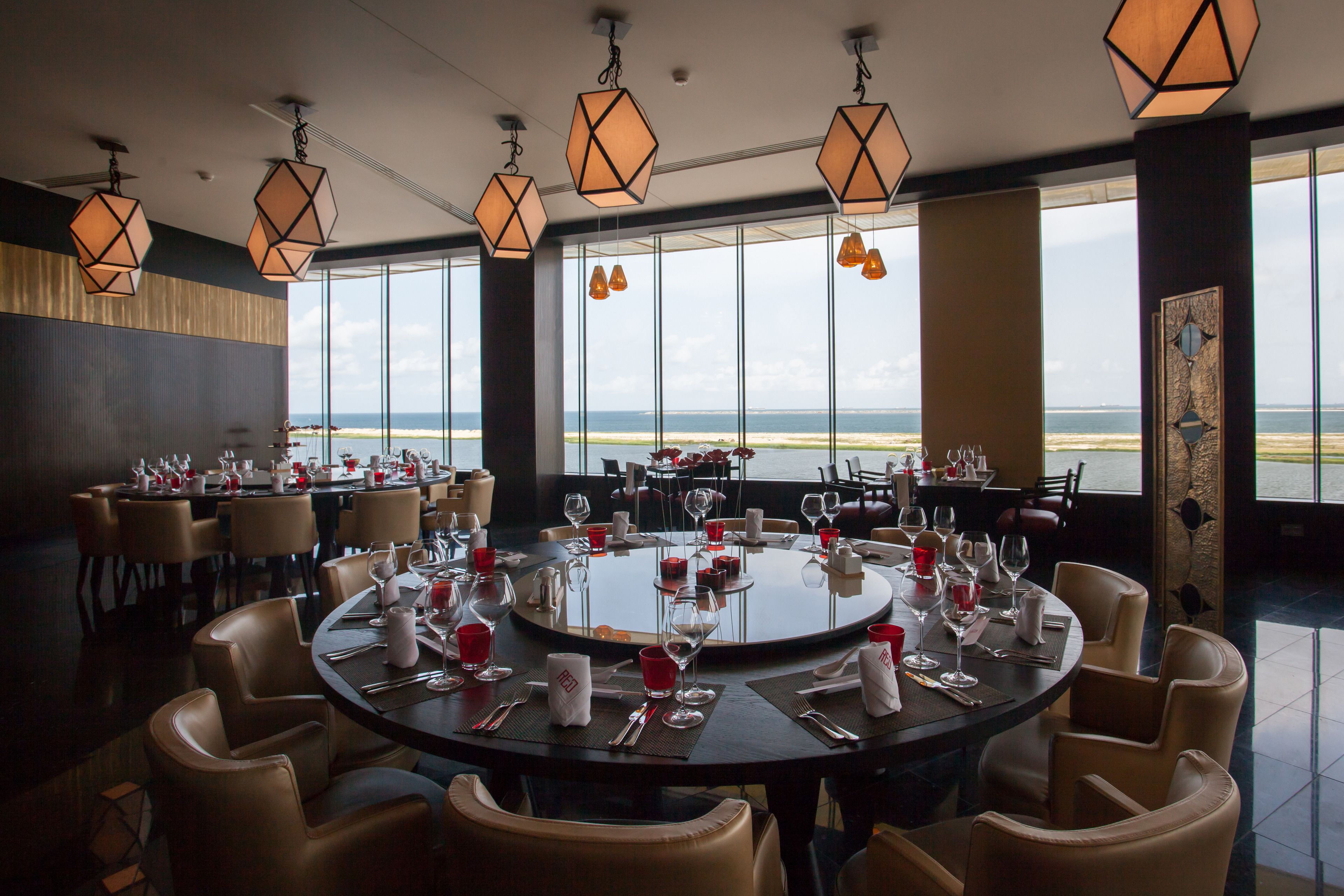 A restaurant with round tables set for dining, large windows with a view of the sea, and geometric pendant lighting.