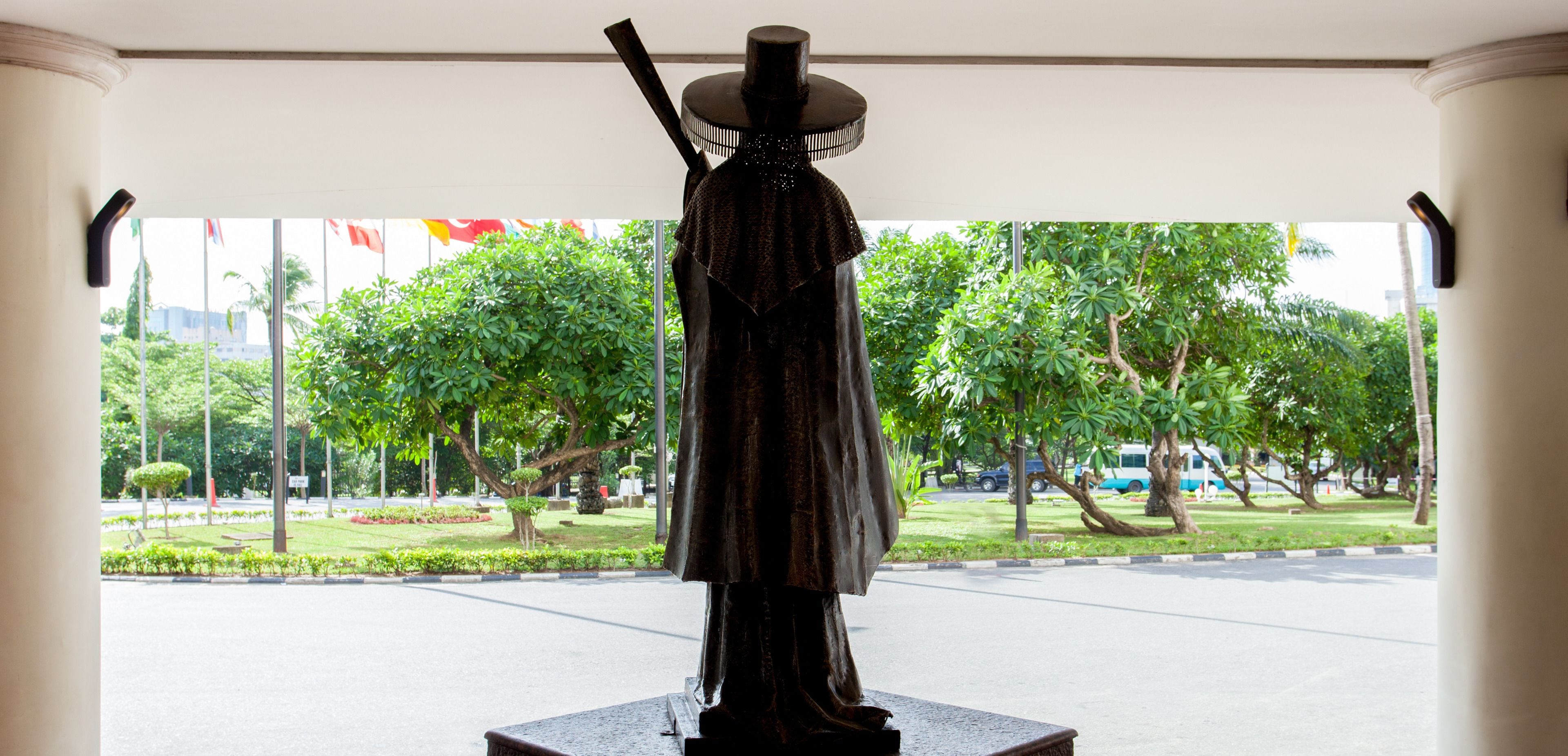 A statue of a person in traditional attire, standing on a pedestal, with a view of a well-maintained garden and a road in the background.