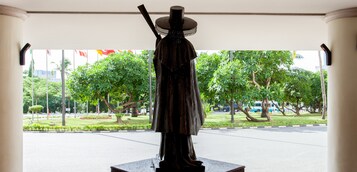 A statue of a person in traditional attire, standing on a pedestal, with a view of a well-maintained garden and a road in the background.