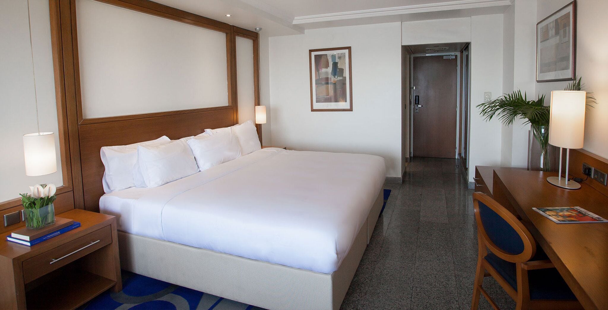 A hotel room with a large bed, a wooden headboard, a desk with a lamp, a chair, and a framed picture on the wall.