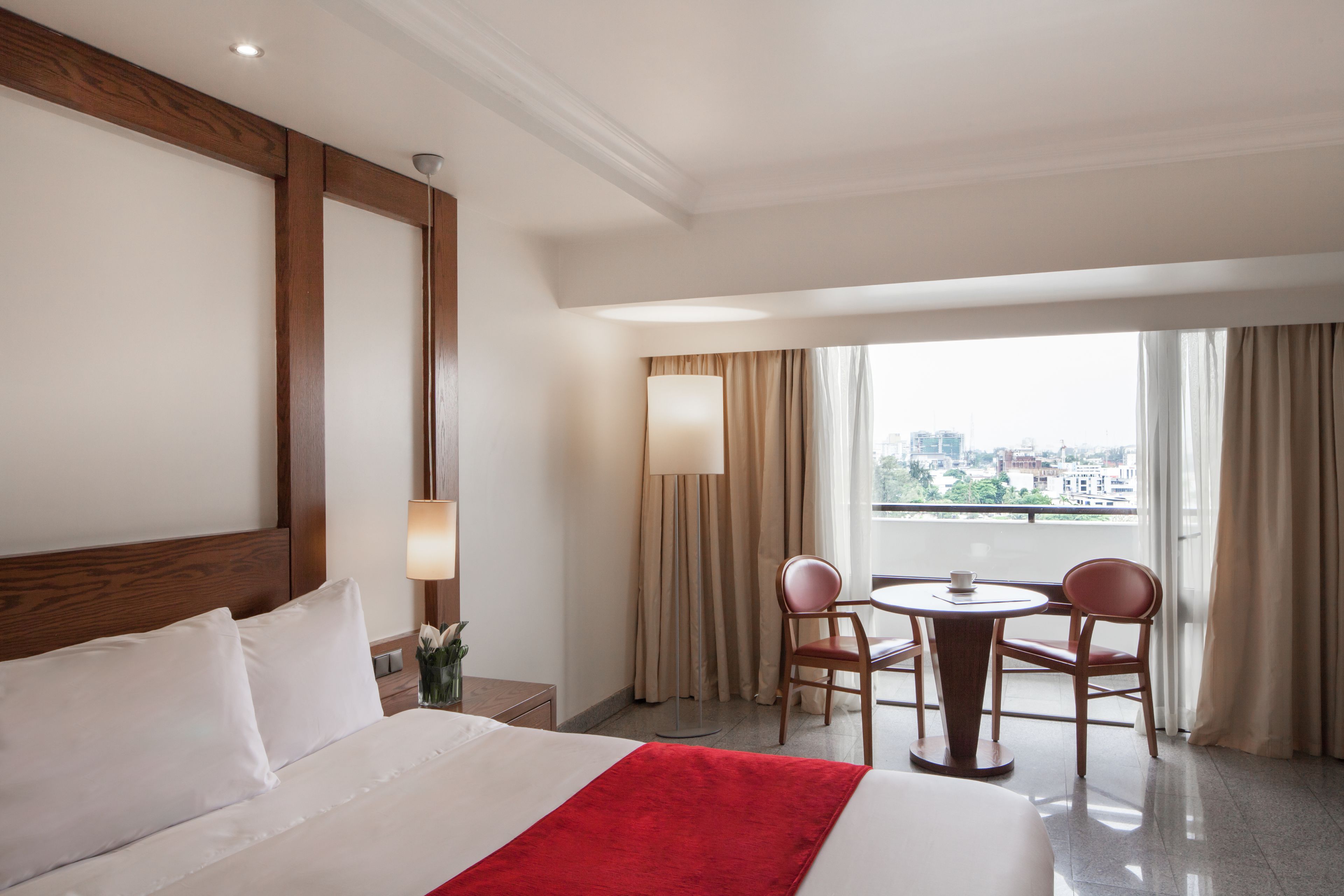 Una camera d'albergo con un letto grande, un tavolino con due sedie, una lampada e vista sulla città attraverso ampie finestre.