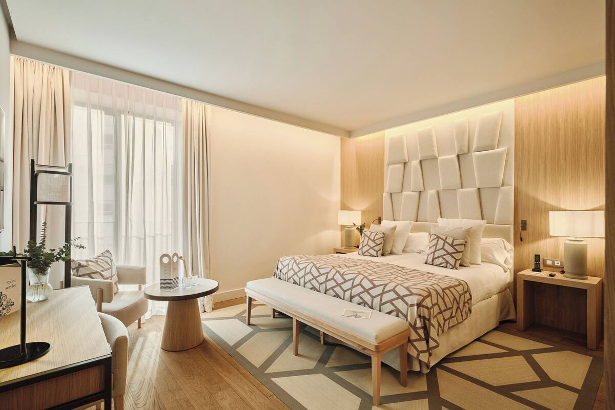 Une chambre d’hôtel moderne, dotée d’un grand lit, de tables de chevet, d’une banquette, d’une petite table ronde et d’un bureau avec une chaise.