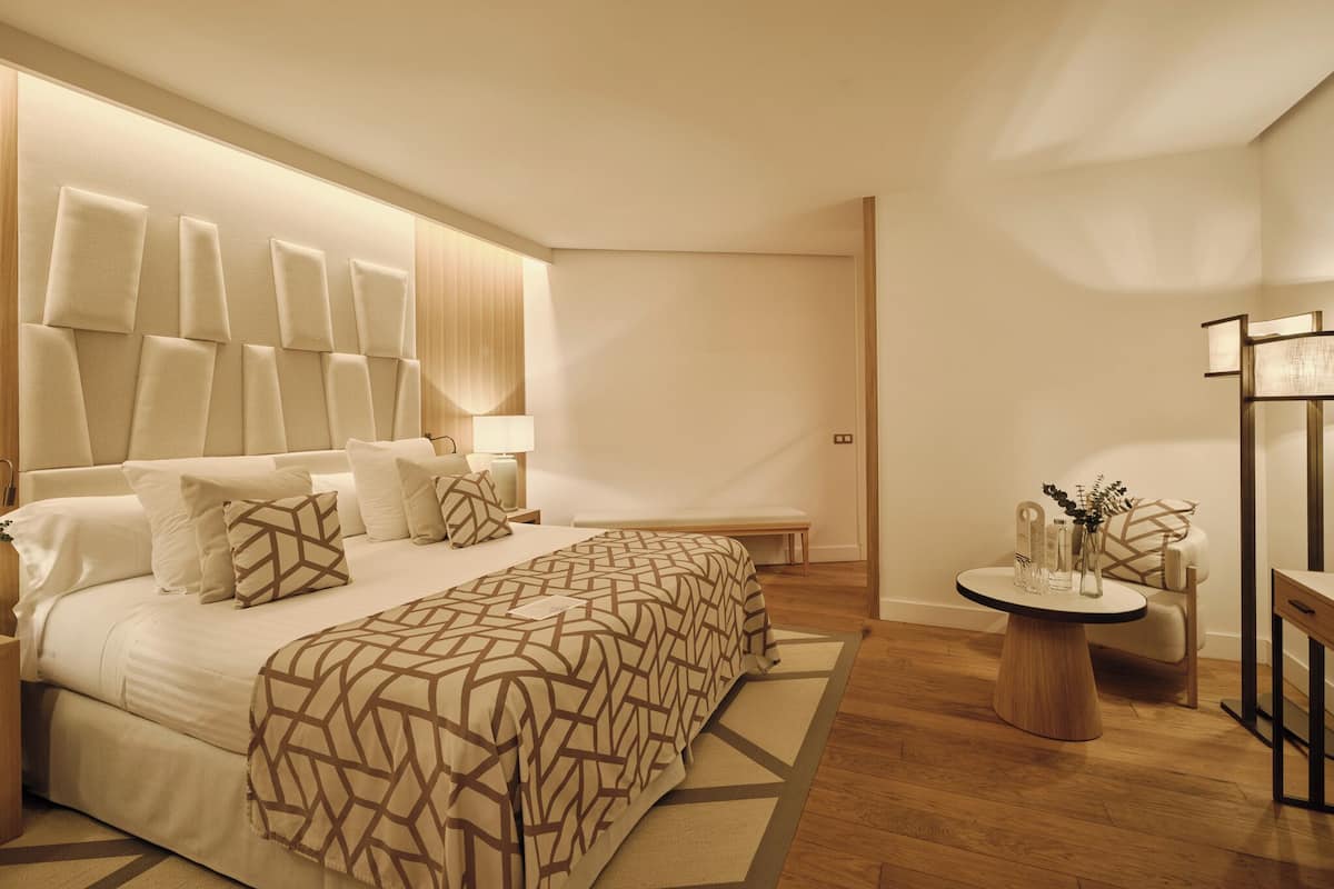 Une chambre à coucher moderne avec un grand lit, des tables de chevet, une petite table d’appoint ronde en bois et une chaise.