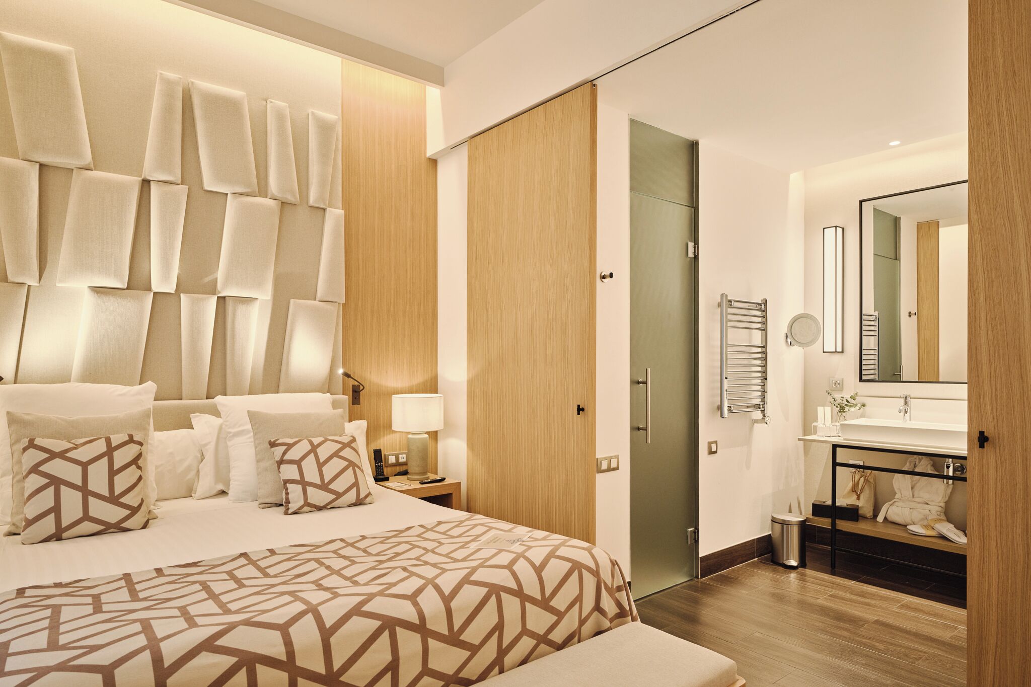 A modern hotel room with a large bed, a wooden wardrobe, a bathroom with a mirror, and a small seating area.