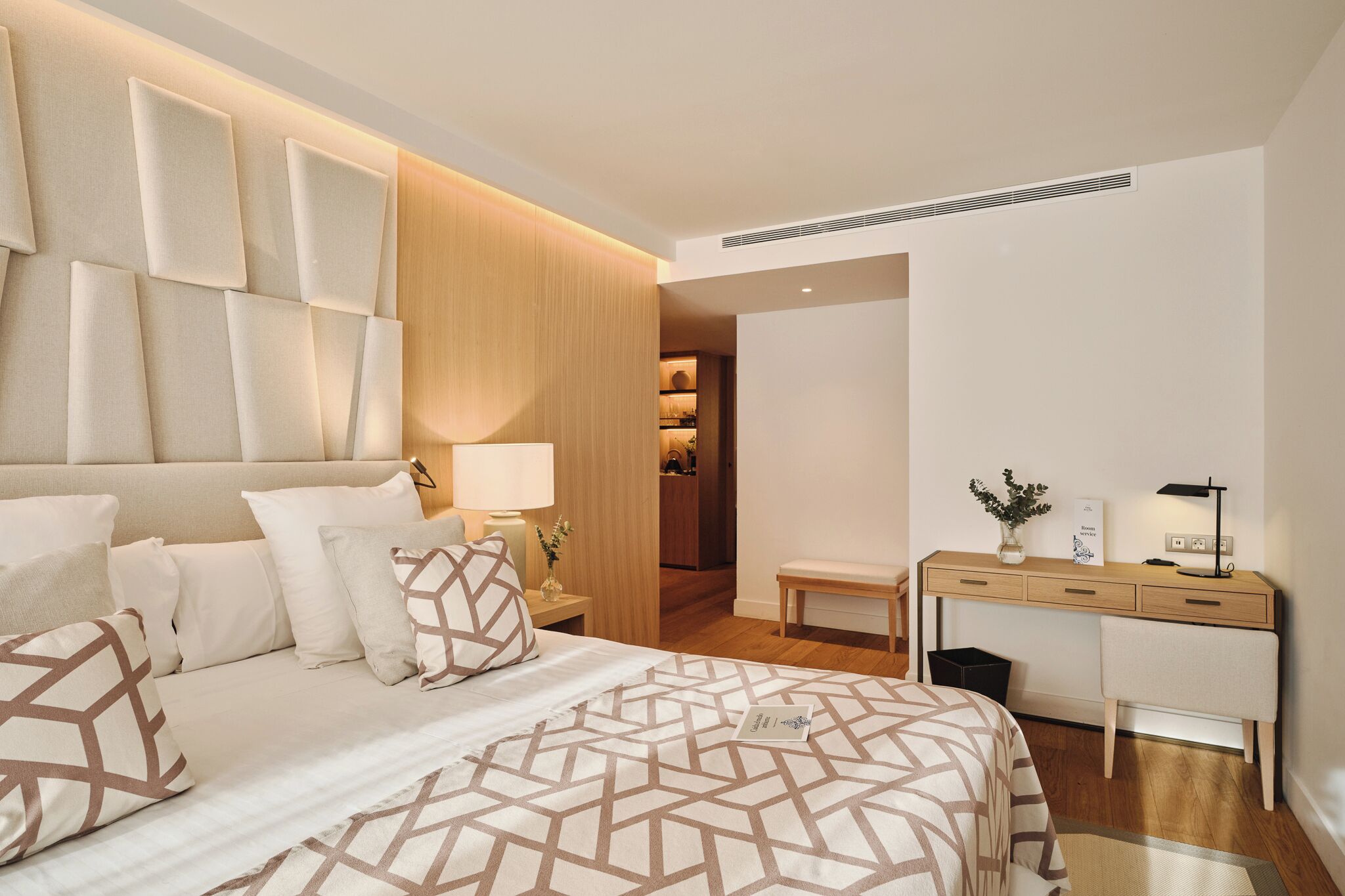 A modern hotel room with a large bed, a wooden headboard, a desk with a lamp, and a small bench.