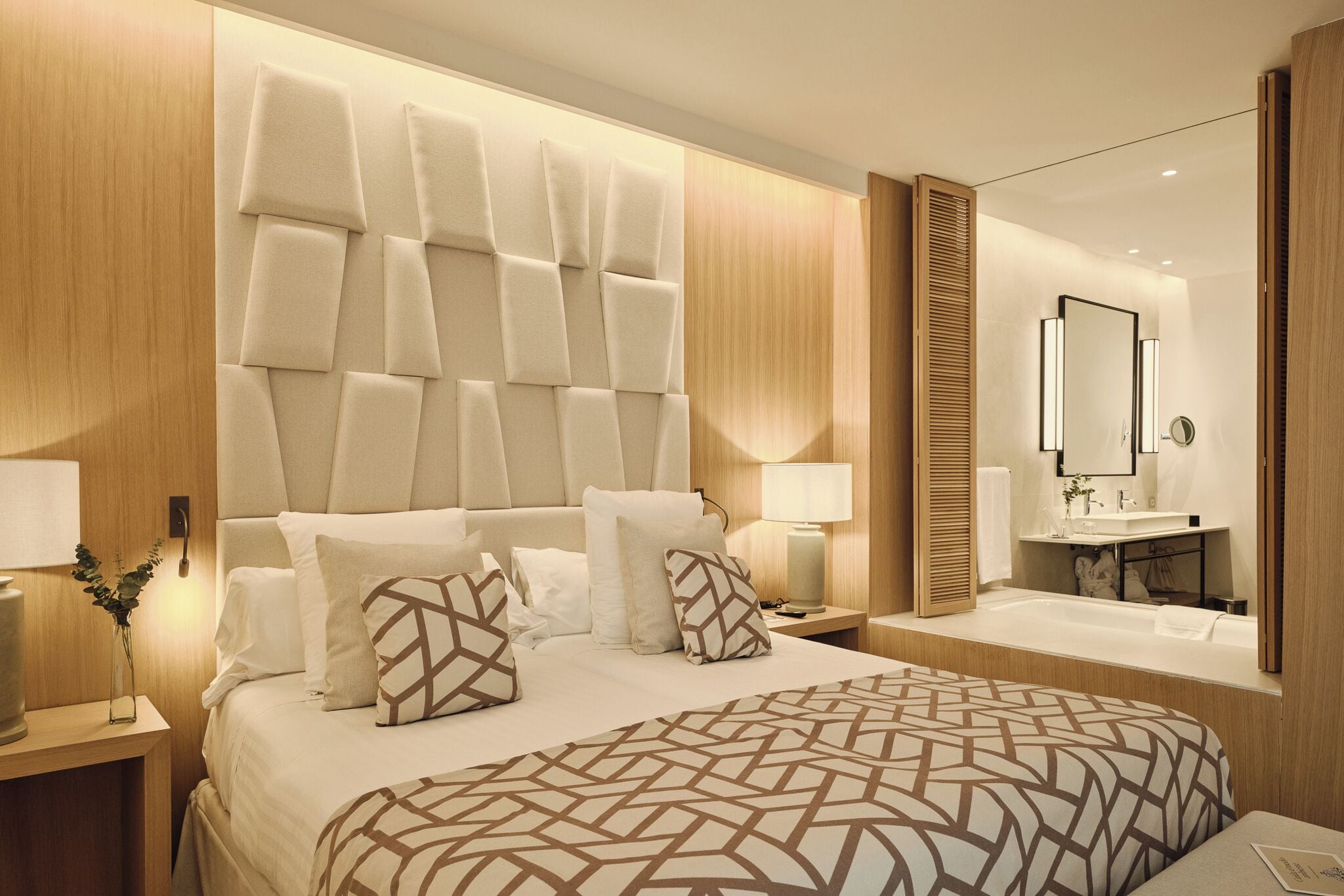A modern hotel room with a large bed, a bedside table with a lamp, a built-in headboard, a mirror, and a bathroom with a sink and bathtub.