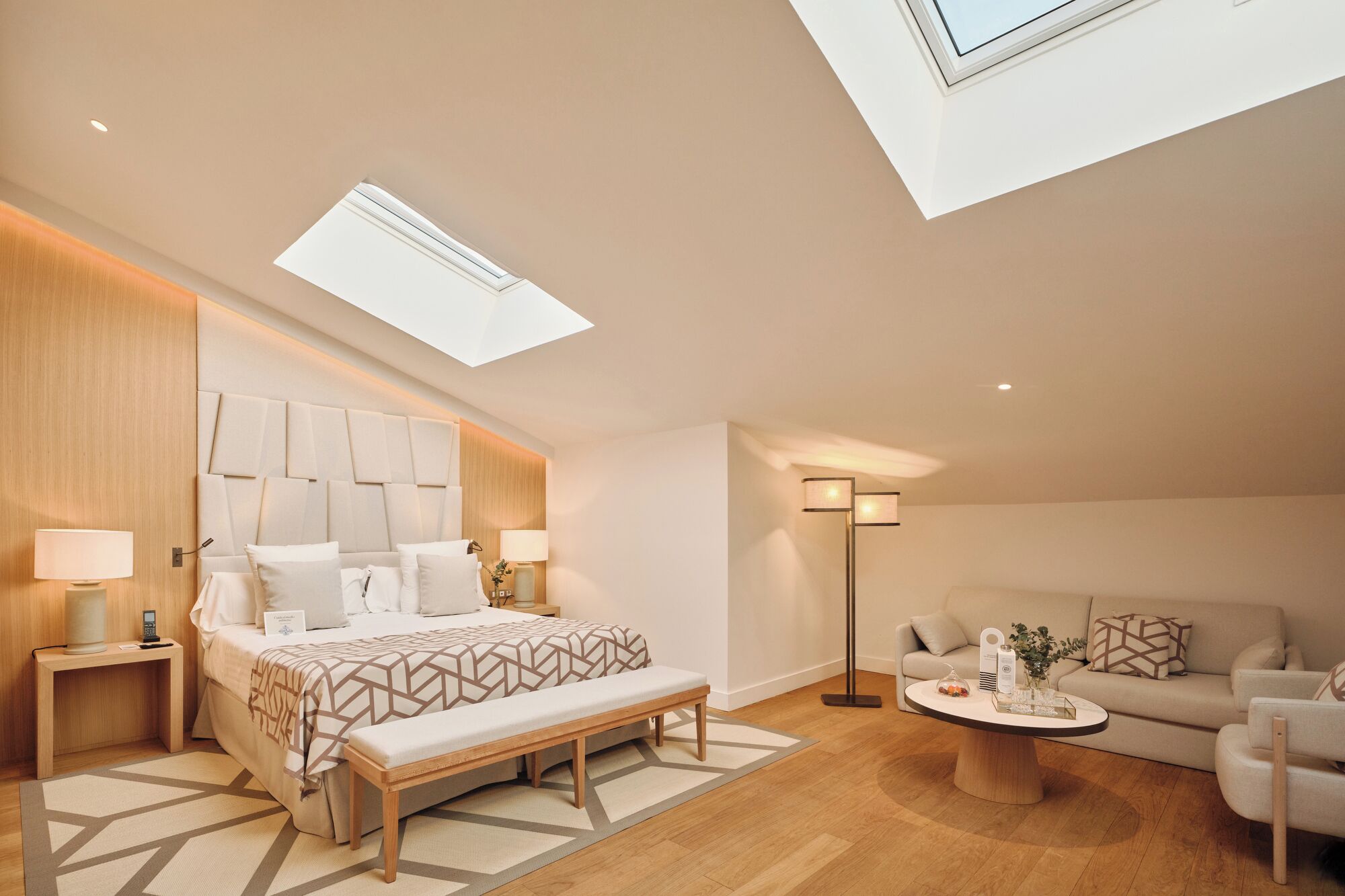 A modern bedroom with a skylight, a bed with a geometric-patterned bedspread, a bedside table with a lamp, a bench, a bedside table with a phone, a floor lamp, a sofa, and a coffee table with decorative items.