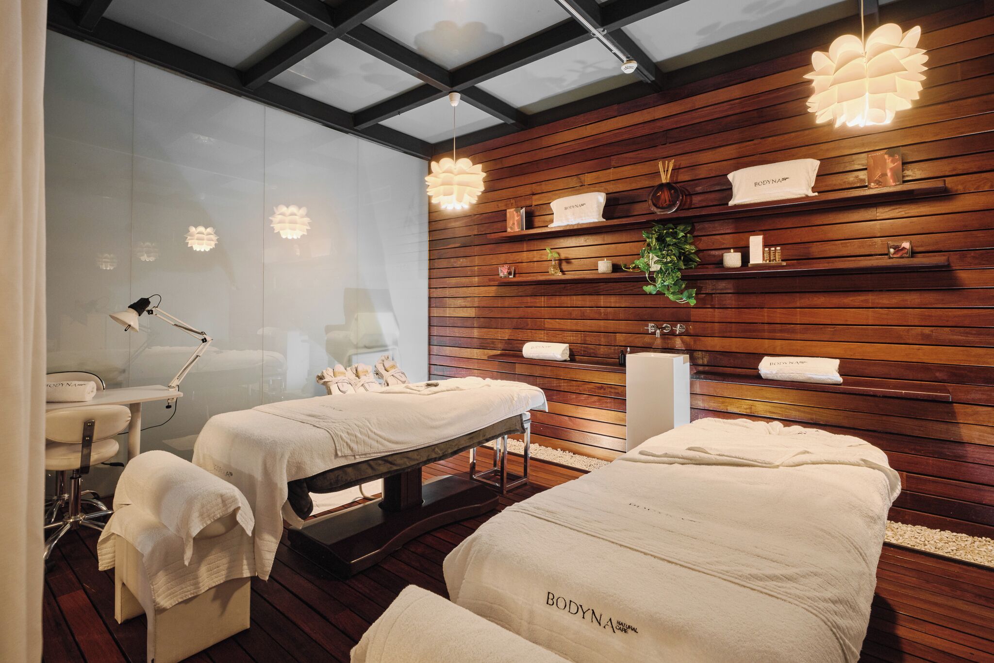 A spa room with wooden paneling, two massage tables, a mirror, and a ceiling light fixture.