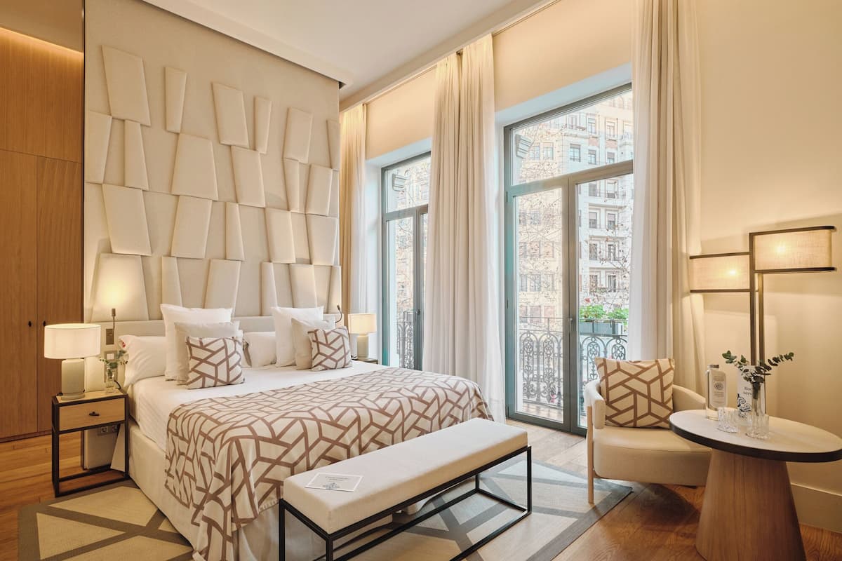 Une chambre d’hôtel moderne, dotée d’un grand lit, d’une banquette, d’une chaise, d’une table et offrant une vue sur la ville grâce à de grandes fenêtres.