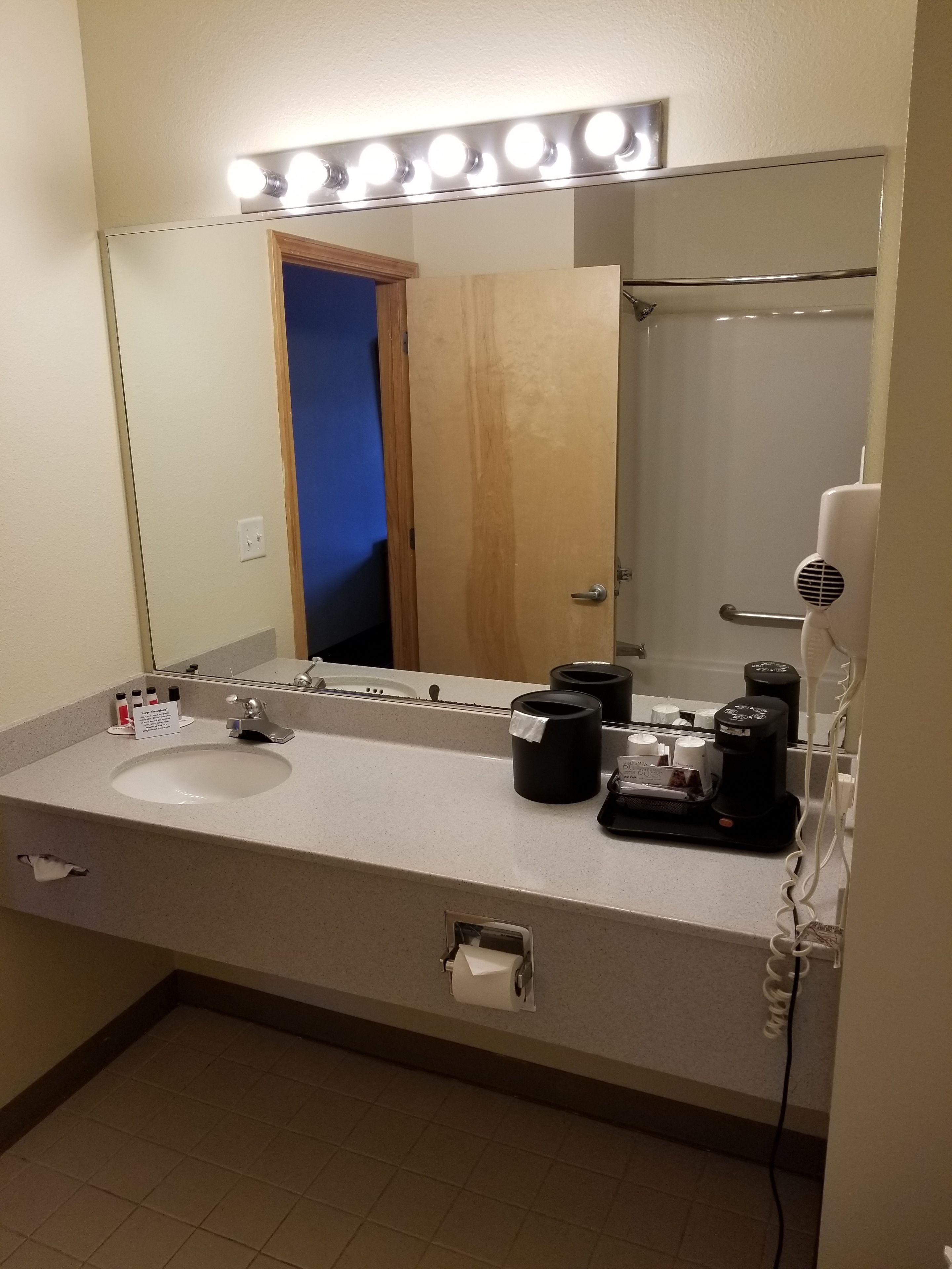 A bathroom with a sink, mirror, and lights above it. There is a towel rack and a hairdryer on the right.