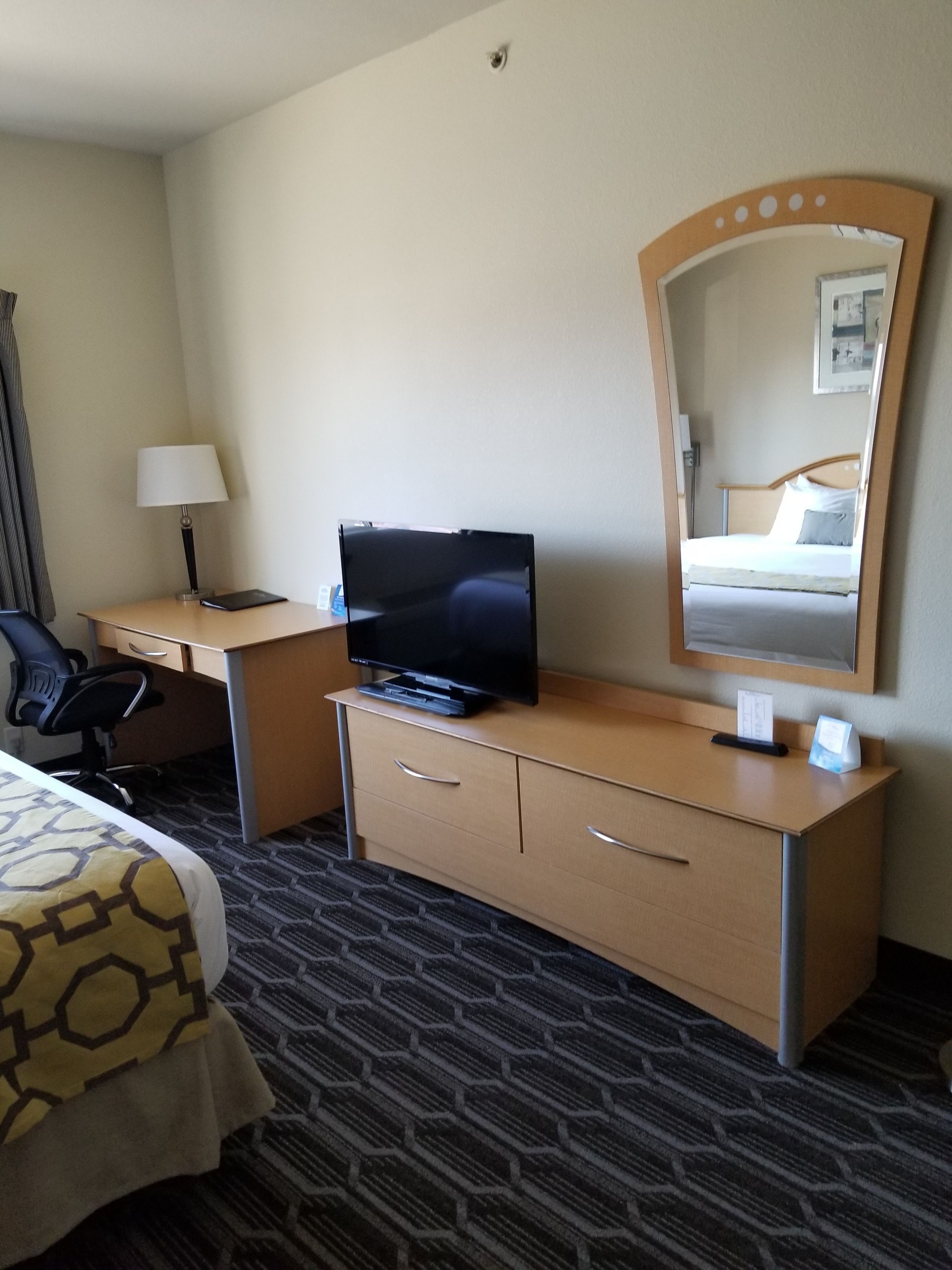 Una camera d'albergo con un letto, una scrivania con televisore, una sedia, una lampada, una cassettiera e uno specchio.