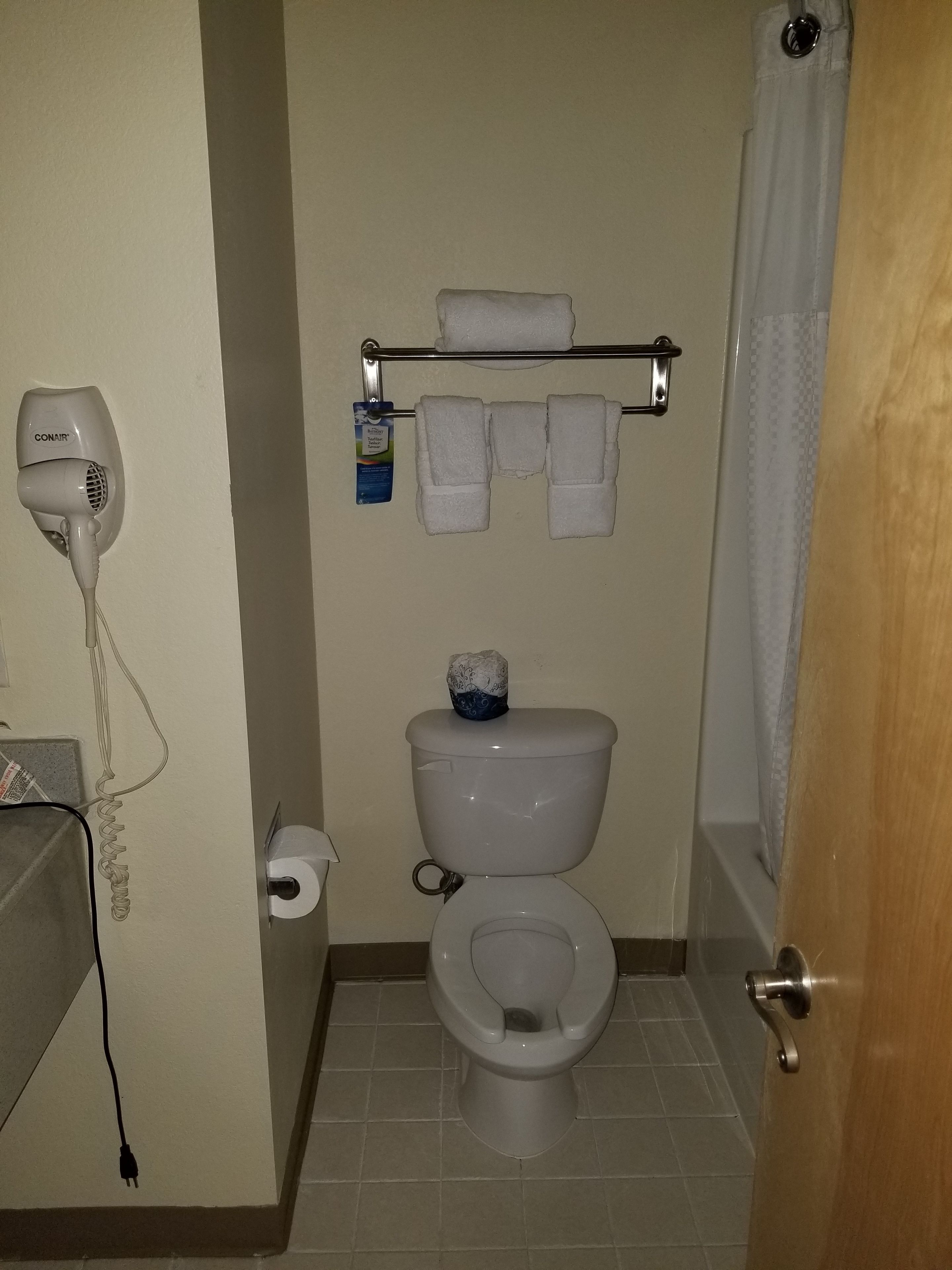 A bathroom with a toilet, a towel rack with towels, a paper towel dispenser, and a hair dryer plugged in.