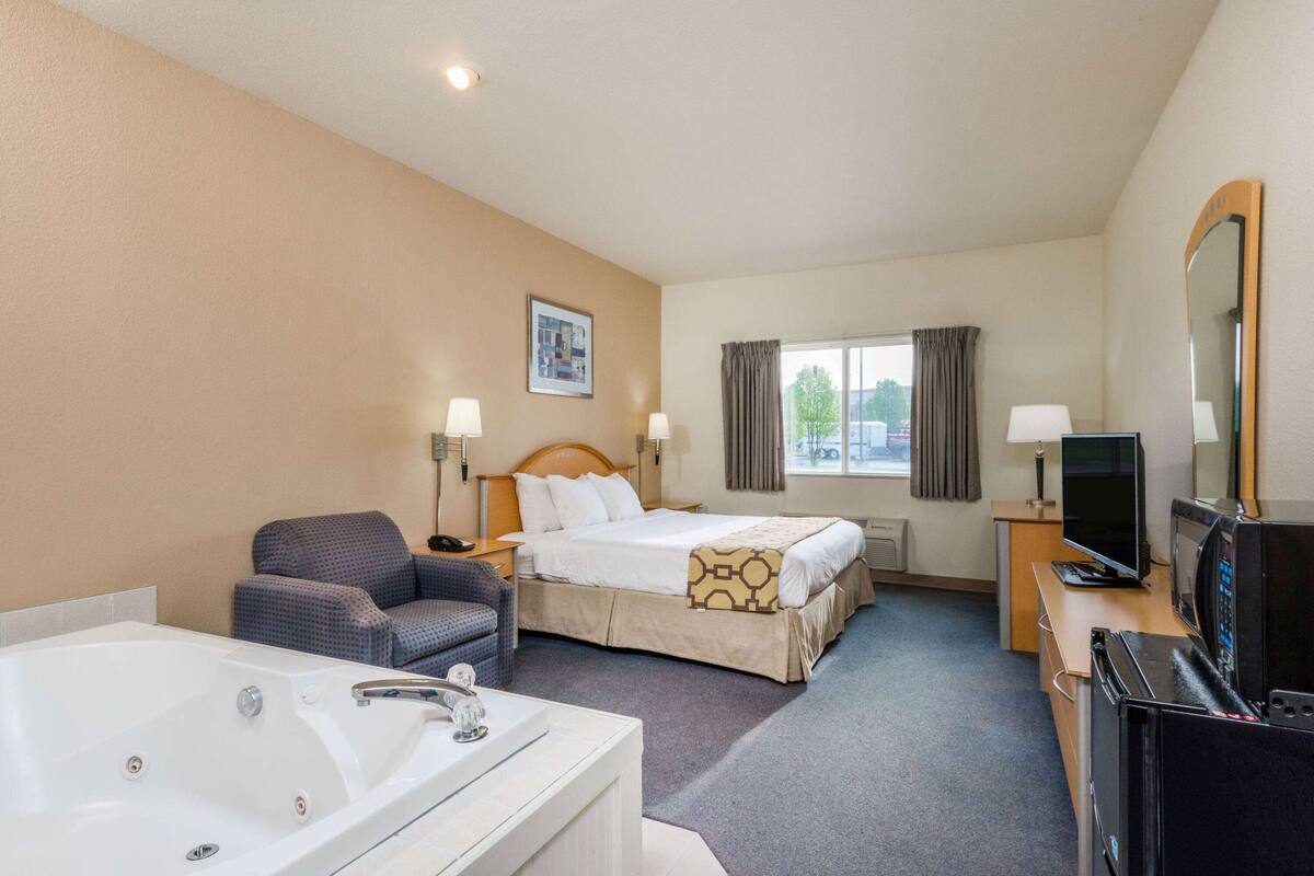 Ein Hotelzimmer mit einem großen Bett, einer Sitzecke mit einem Sessel, einem Schreibtisch mit Computer, einem Fernseher und einer Badewanne.