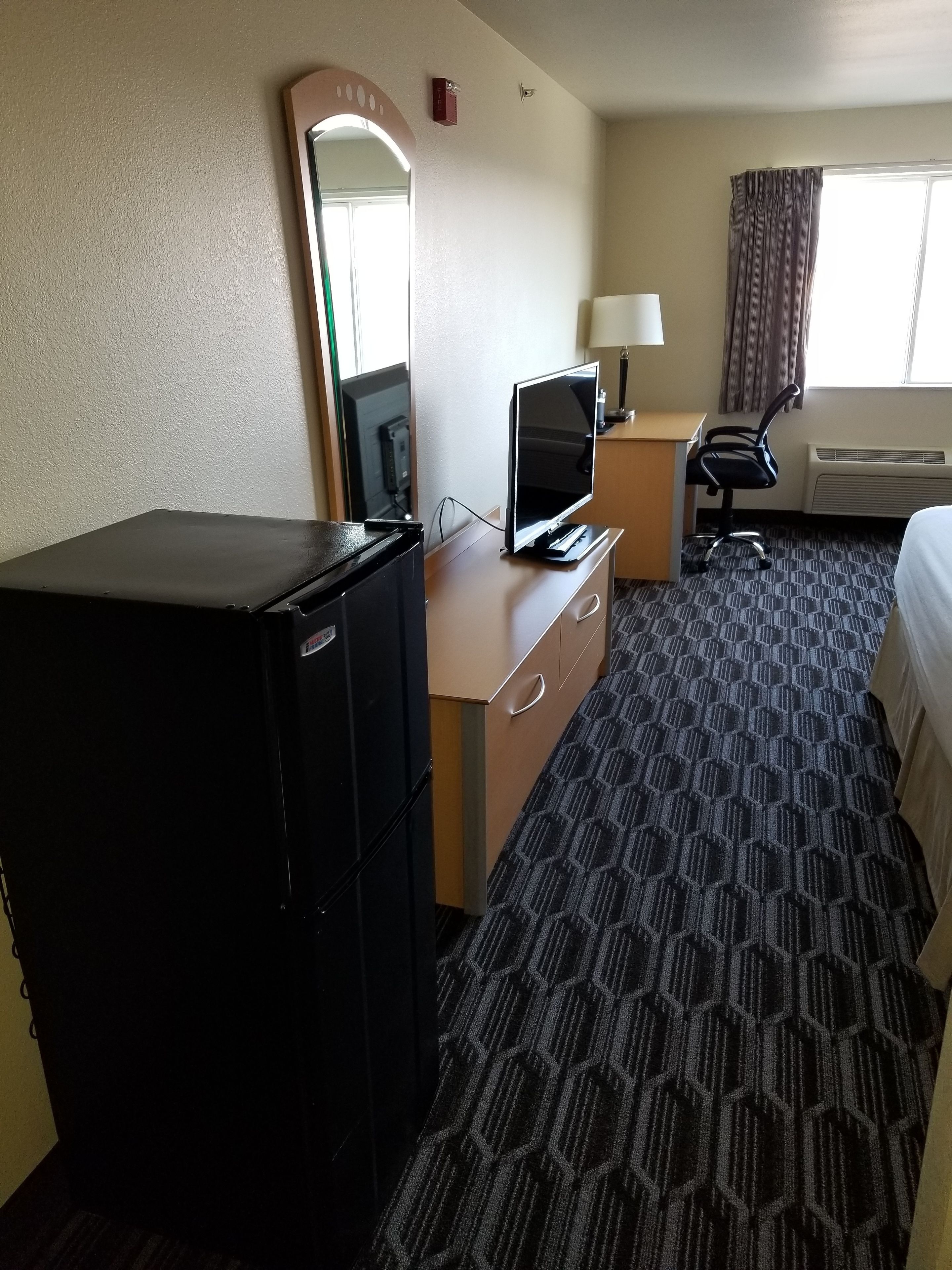 A hotel room with a bed, a desk with a TV, a dresser, a mirror, a lamp, and an air conditioning unit.