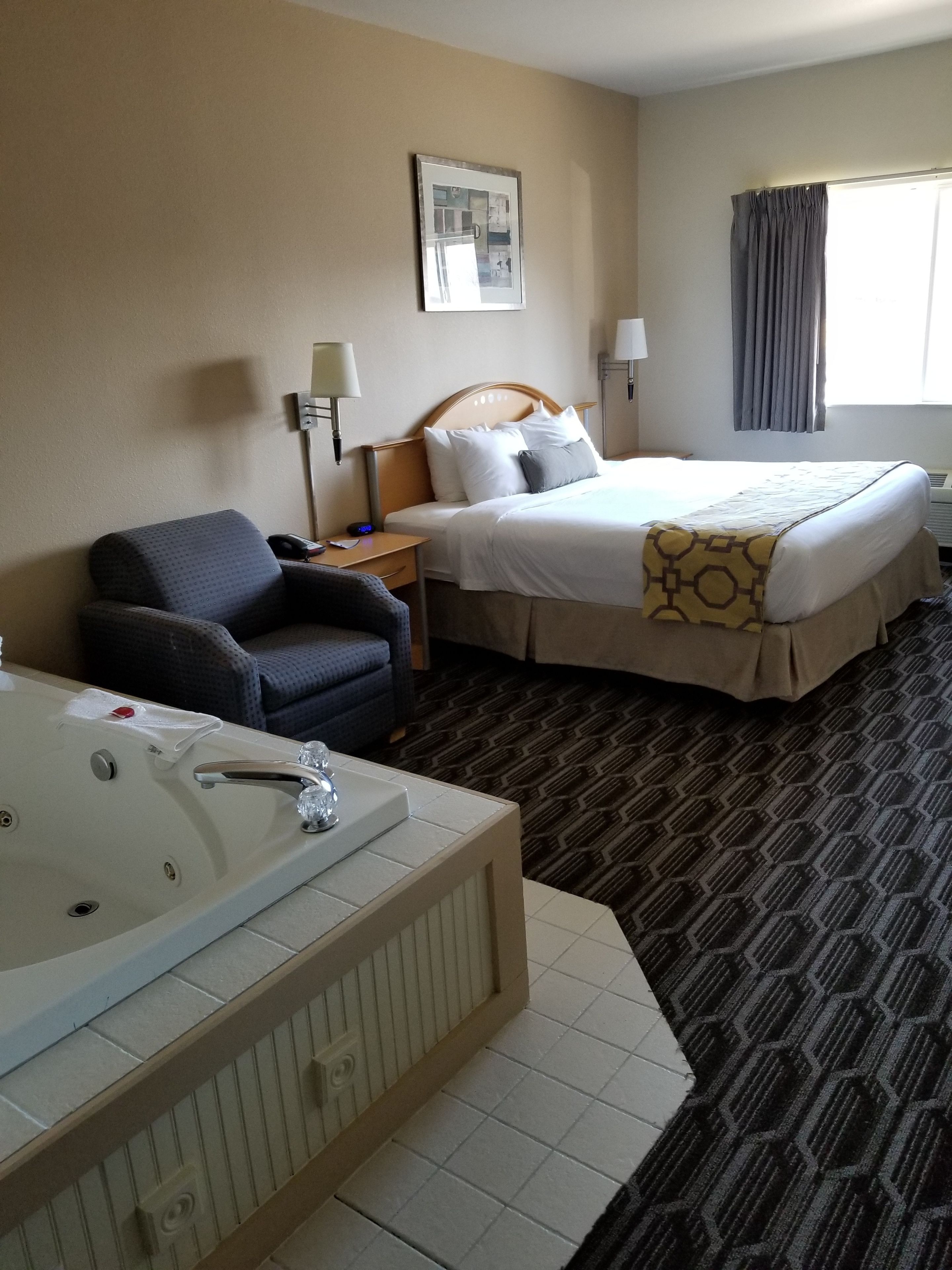 A hotel room with a bed, a chair, a desk, a lamp, a picture on the wall, and a window with curtains.