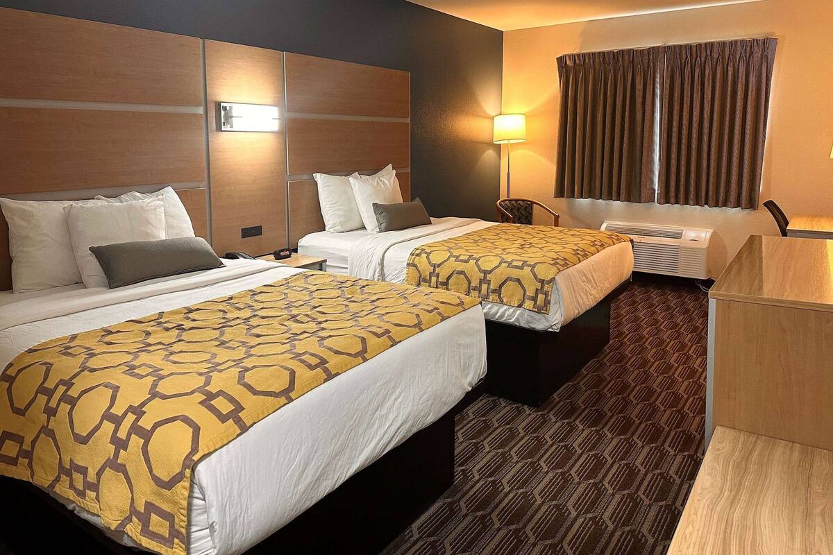 Ein Hotelzimmer mit zwei Betten, einem Schreibtisch, einem Stuhl, einer Lampe und einem Fenster mit Vorhängen.