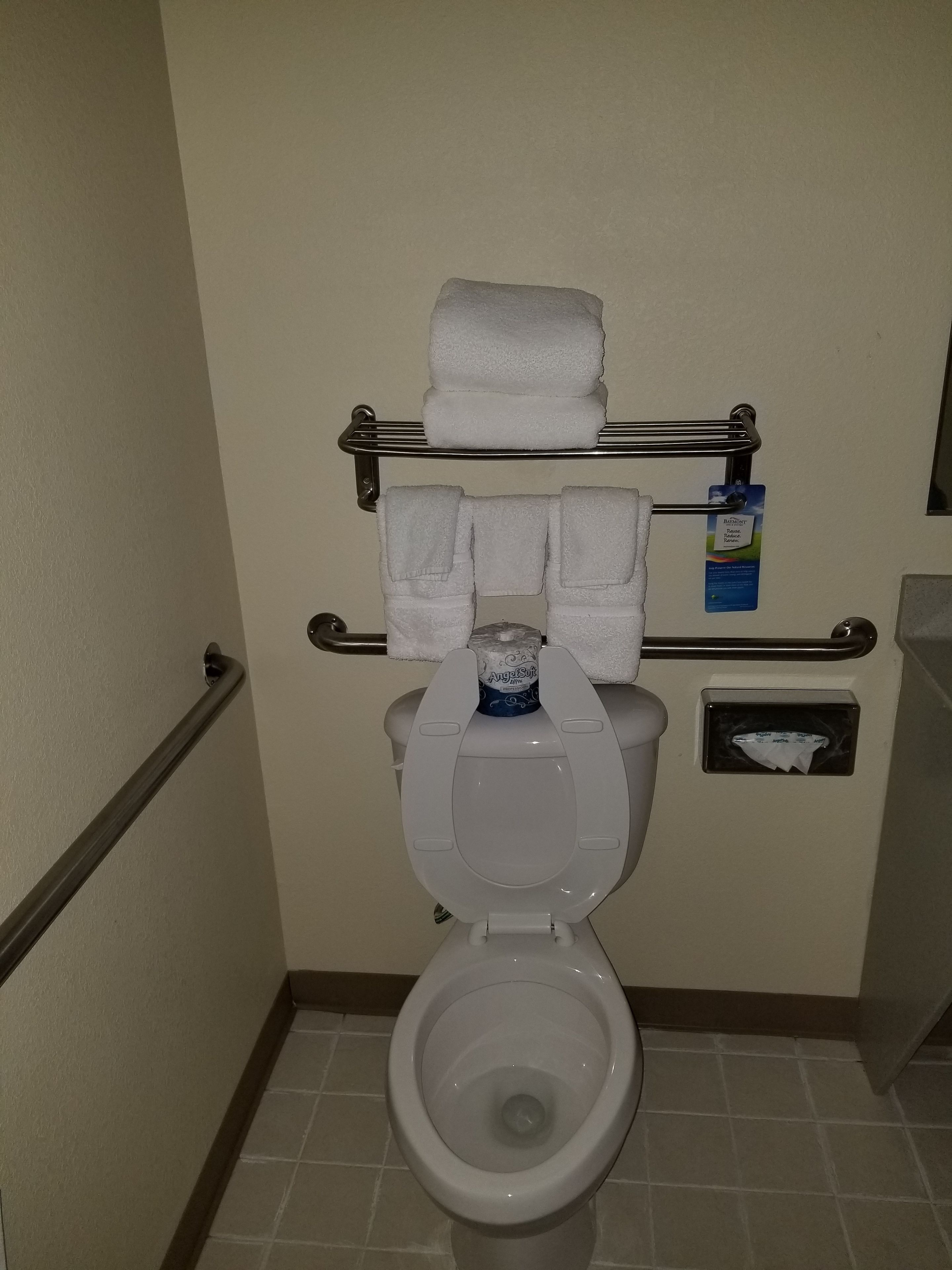A bathroom with a toilet, a towel rack with folded towels, a toilet paper holder, and a hand sanitizer dispenser.