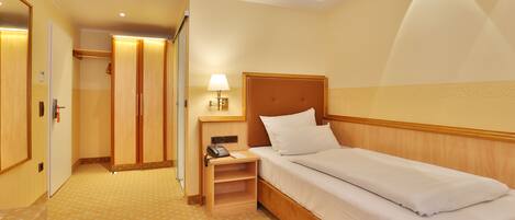 Quarto de hotel com uma cama de solteiro, mesa de cabeceira com abajur, telefone e guarda-roupa.