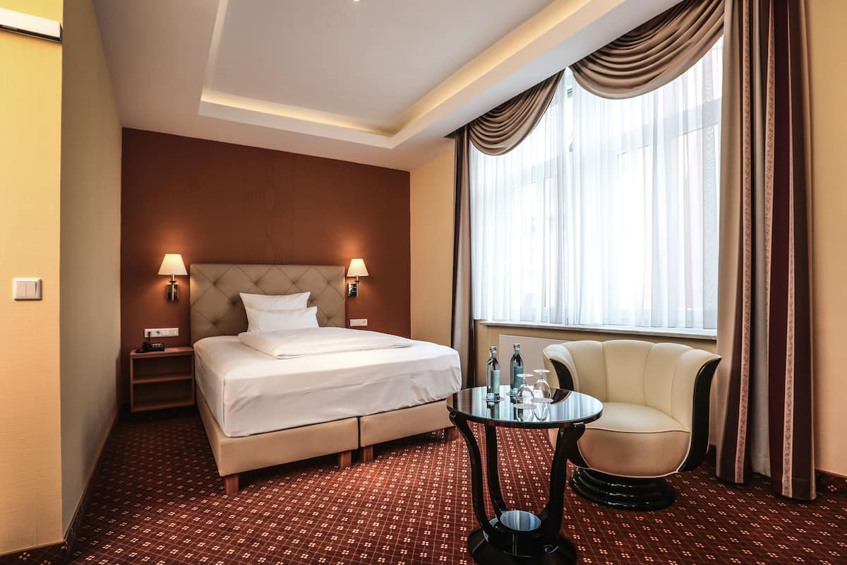 A hotel room with a bed, bedside lamps, a round table, a chair, and a large window with curtains.