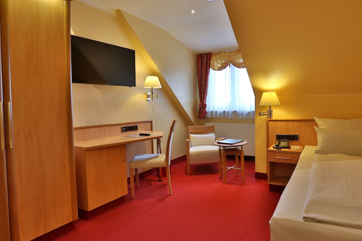 A hotel room with a bed, a desk with a chair, a small table with a chair, a TV, and a window with curtains.