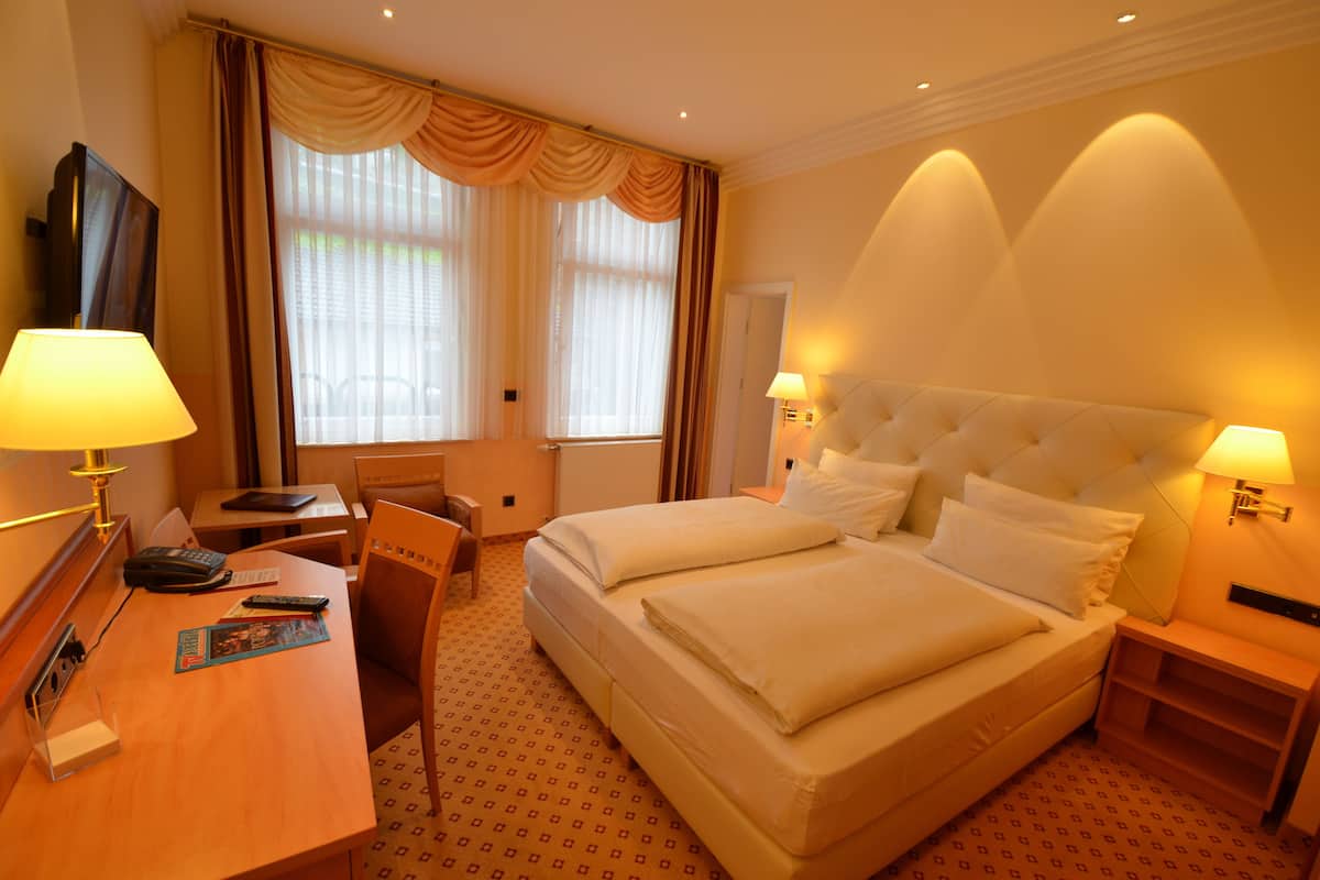 A hotel room with a large bed, two bedside tables with lamps, a desk with a chair, a TV, and a window with curtains.