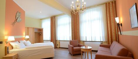 A hotel room with a bed, two armchairs, a small table, a chandelier, and a view of the outdoors through large windows.