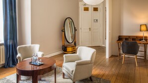A room with a wooden floor, a round wooden table, two white armchairs, a mirror, a lamp, and a door in the background. - Hotel Art Nouveau (Berlin)