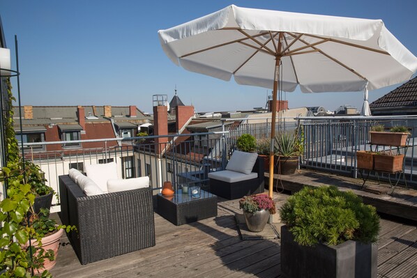 Una terraza en la azotea con mobiliario exterior, una sombrilla grande y plantas en macetas.