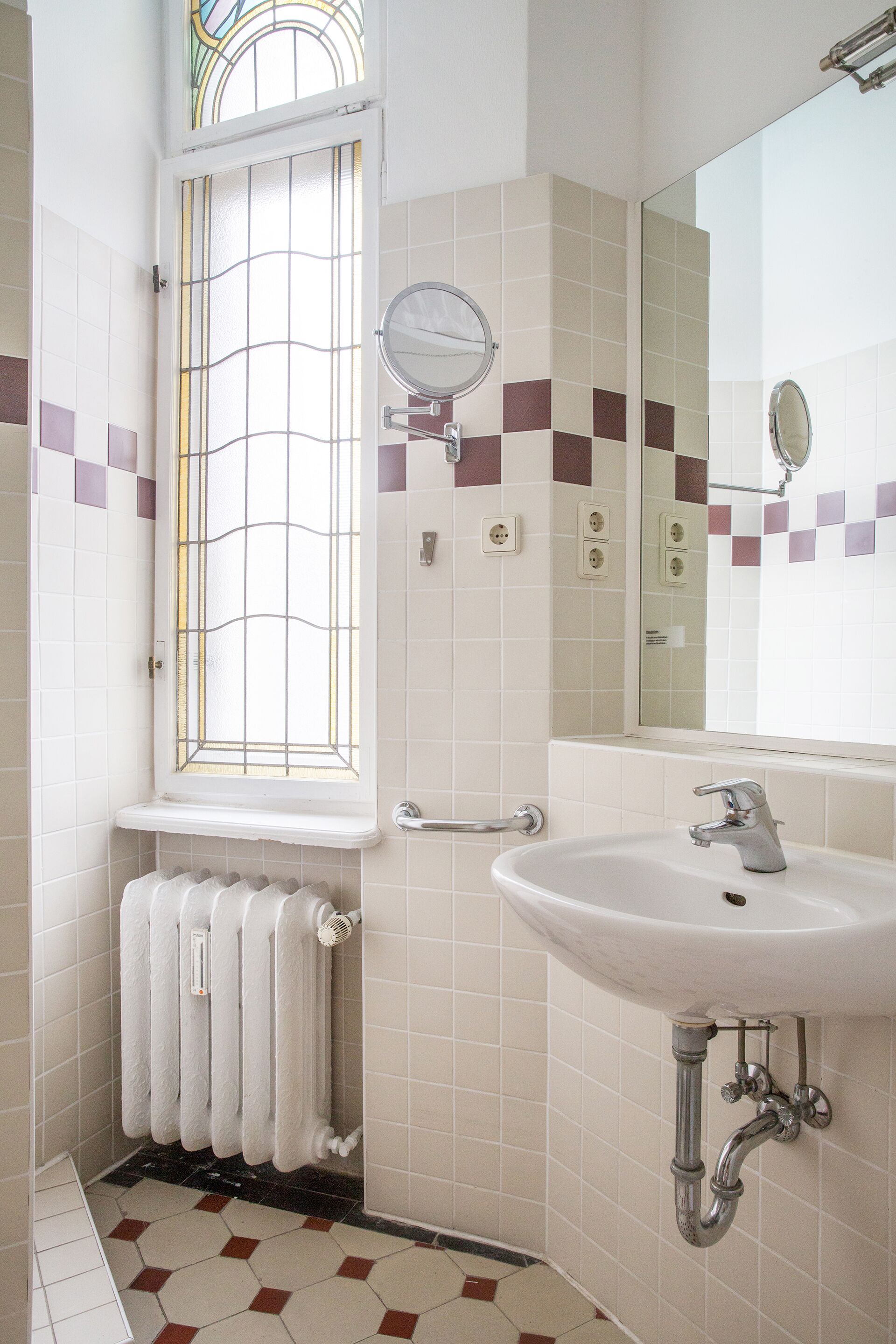 Une salle de bain avec un lavabo blanc, un miroir, une fenêtre ornée de vitraux et un radiateur.