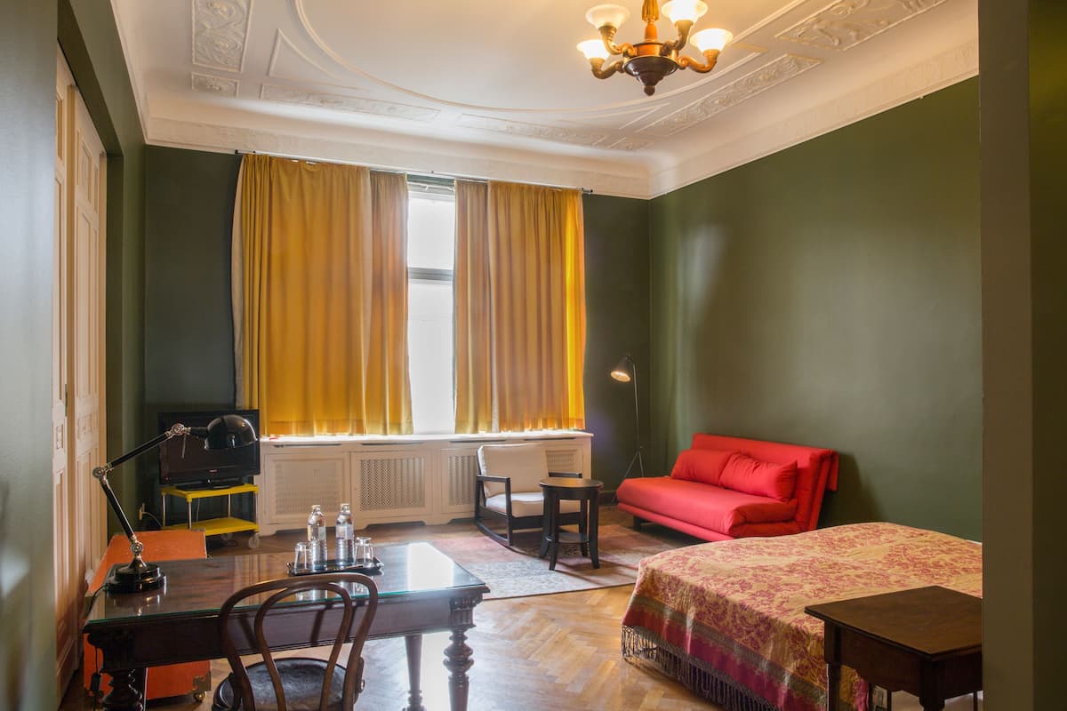 Et hotelværelse med en seng, et skrivebord med stol, en rød sofa, et fjernsyn på et stativ og et vindue med gule gardiner.