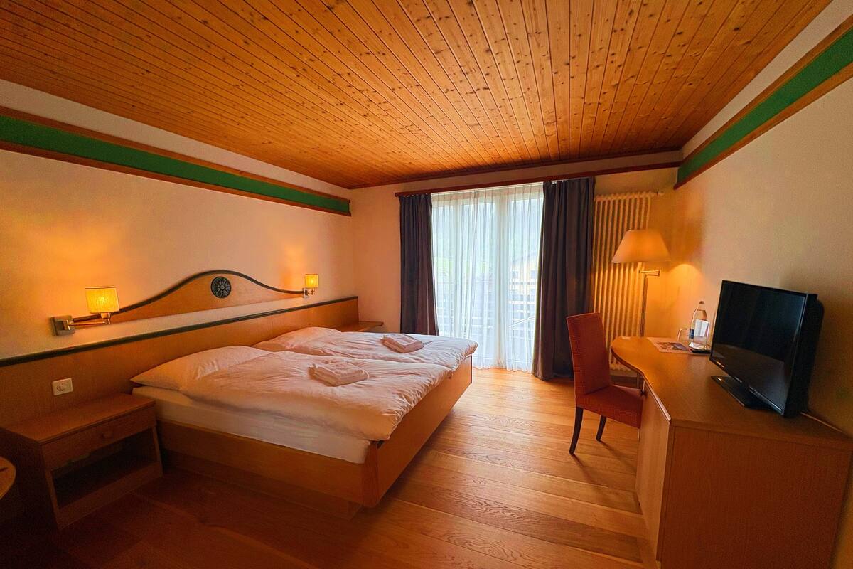 A hotel room with a wooden ceiling, a bed with towels, a desk with a TV, and a chair.