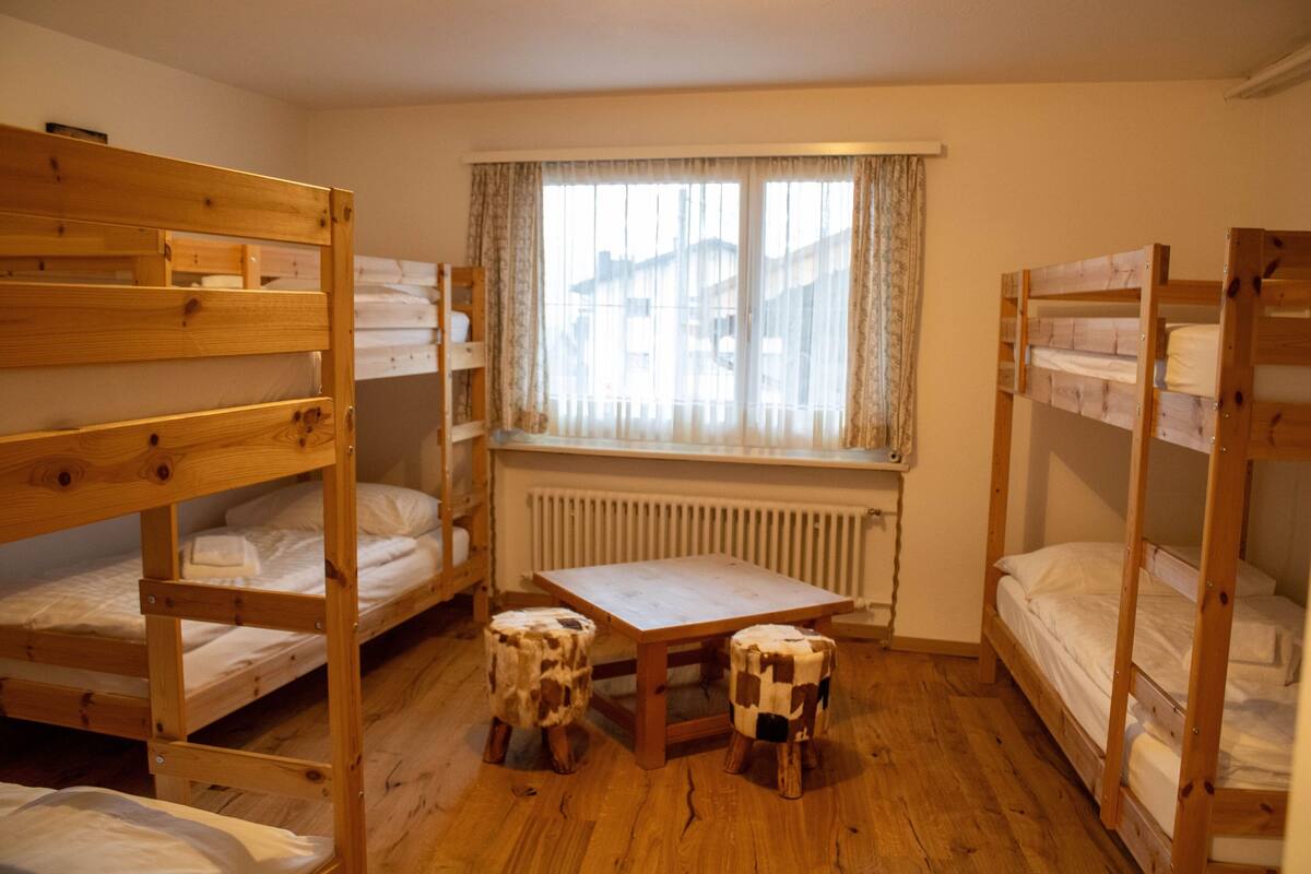 A dormitory room with bunk beds, a small table, two stools, and a window with curtains.