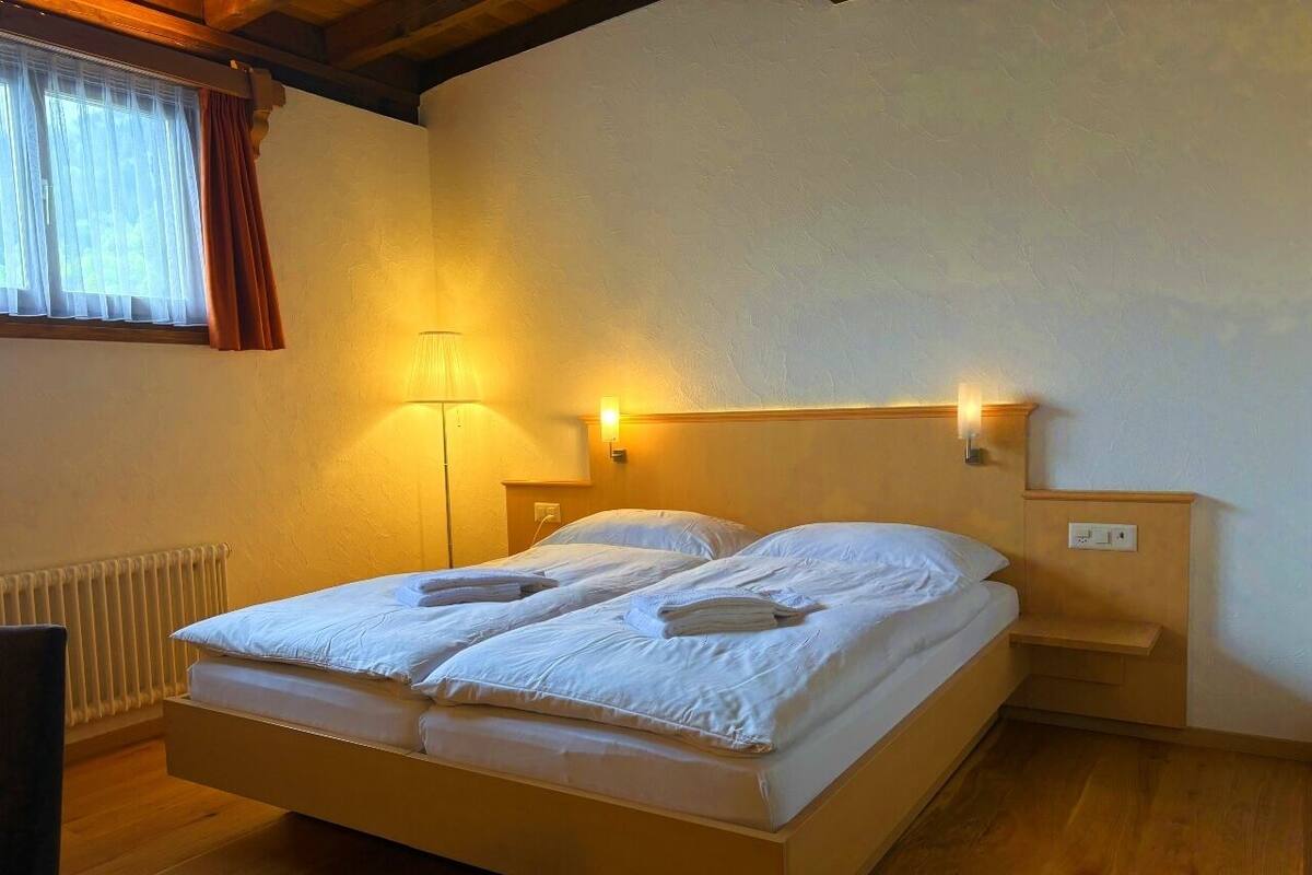 A single bed with white linens, a bedside table, a lamp, and a window with curtains.