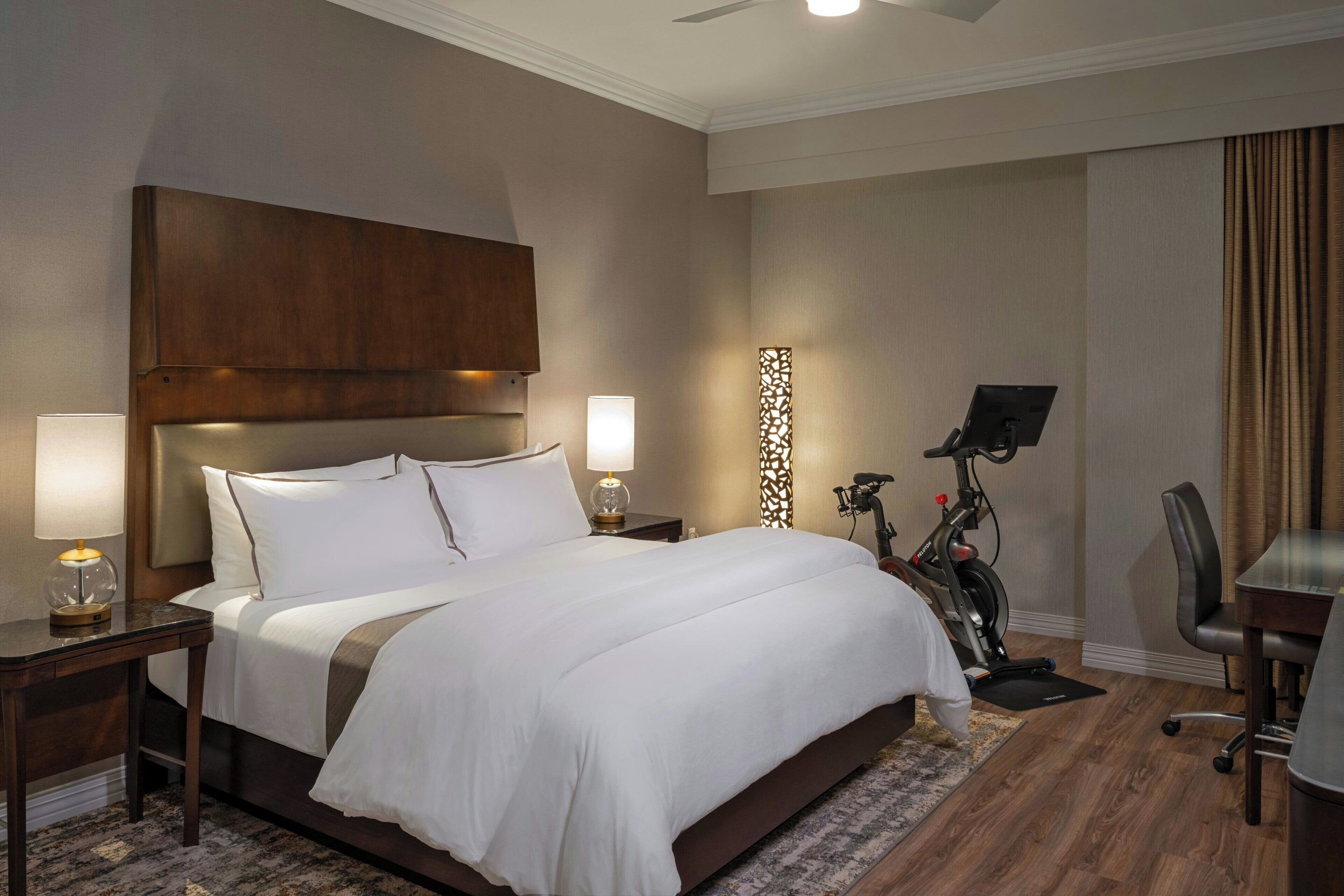 Quarto de hotel com uma cama grande, duas mesas de cabeceira com abajures, uma escrivaninha com cadeira e um ventilador de teto.