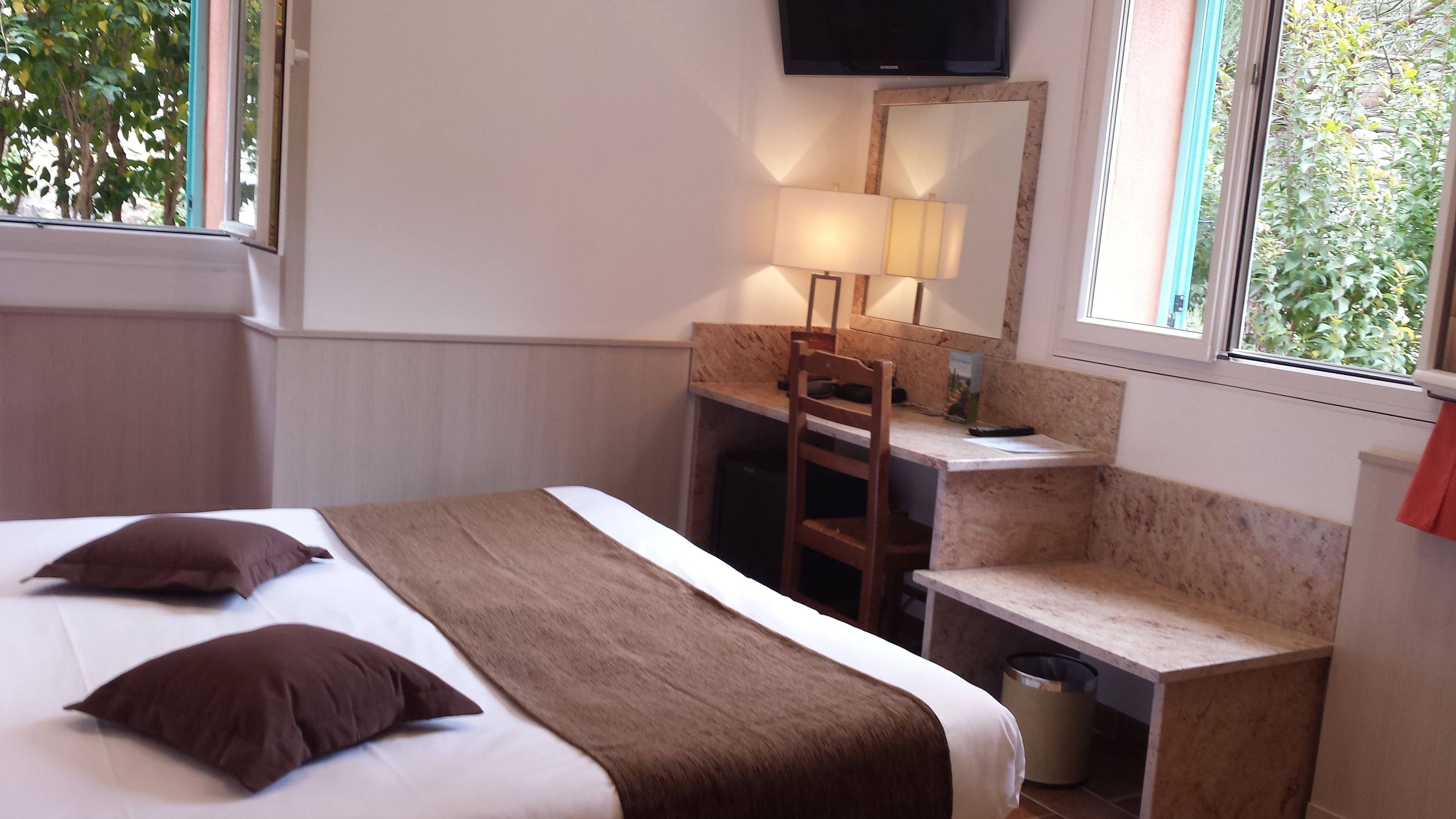 Quarto de hotel com cama, escrivaninha, cadeira, televisão e janela com vista para o verde.