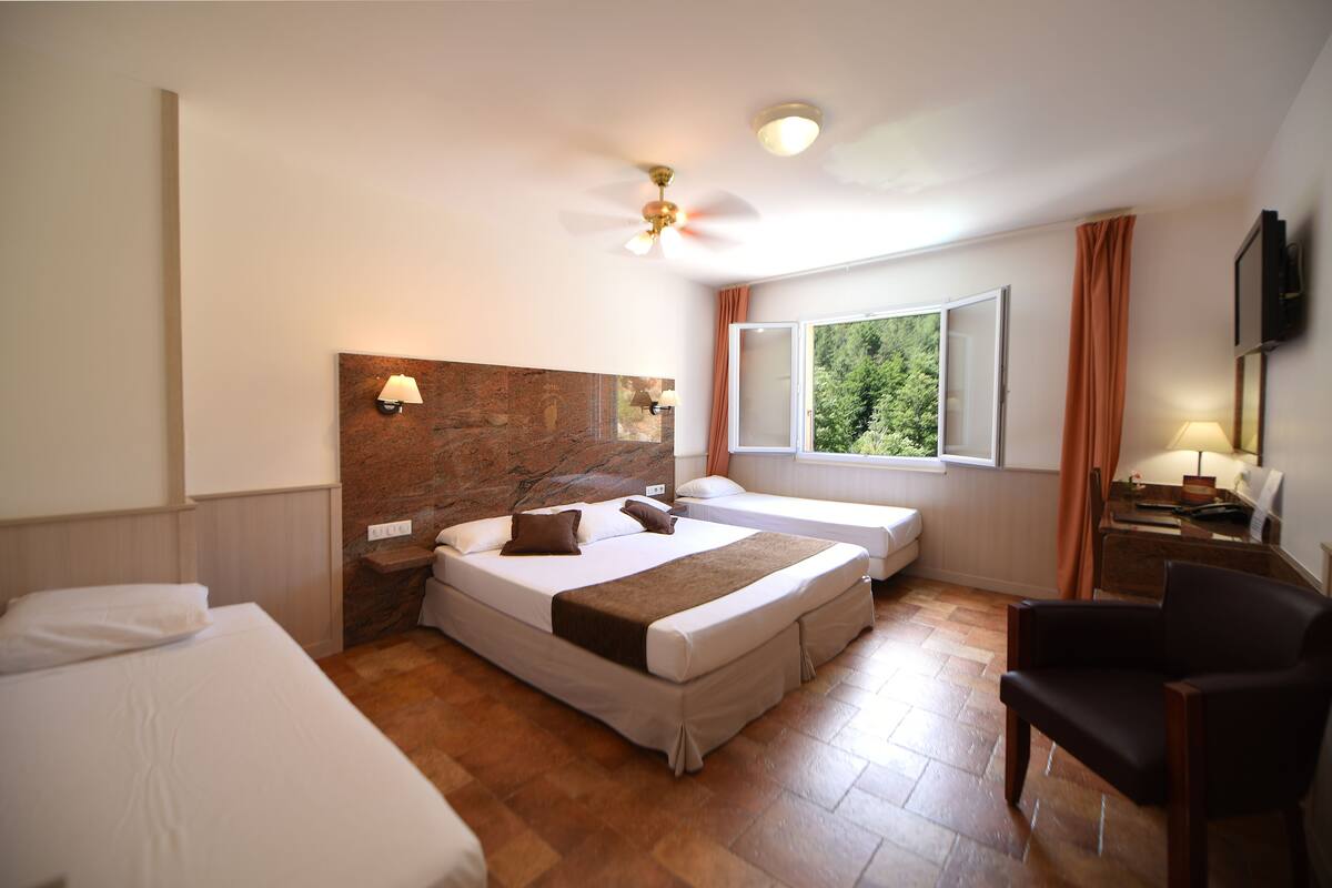 Habitación de hotel con una cama grande, una cama más pequeña, un escritorio con una silla, una ventana con vista a árboles y un ventilador de techo.