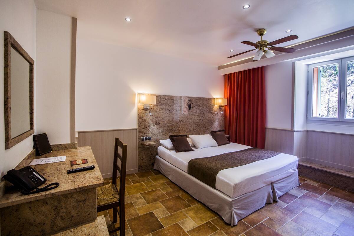 Habitación de hotel con cama, escritorio con teléfono y control remoto, una silla, pared decorativa de piedra, ventilador de techo y ventana con cortinas.