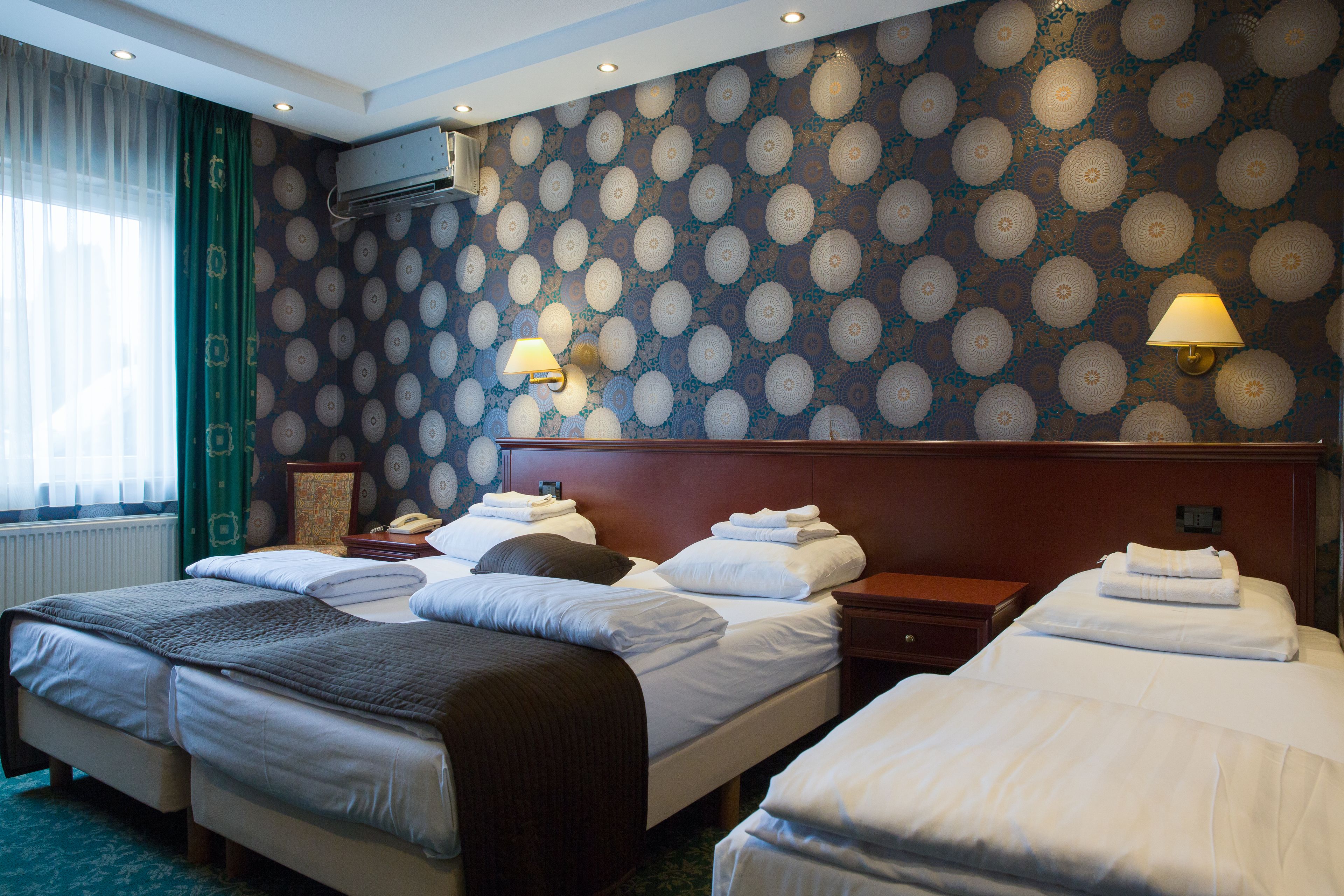 Une chambre d’hôtel avec deux lits, une grande fenêtre avec des rideaux, un papier peint à motifs et deux lampes fixées au mur.