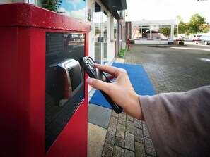 A hand using a mobile device to interact with a red and black automated parking payment kiosk. - Hotel Zwanenburg (Zwanenburg)