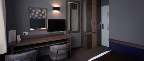 A hotel room with a bed, a desk with two chairs, a flat-screen TV, a large mirror, and a door.